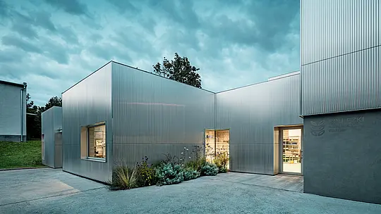 Za súmraku vyniká jedinečný architektonický charakter Envirocentra v Nitre. Hliníková fasáda z vlnitého profilu PREFA v prírodnom hliníkovom vyhotovení sa striebristo trblieta pod dramatickou oblohou plnou oblakov. Vertikálne vlnité línie harmonicky prepájajú jednotlivé časti budovy rôznych výšok, zatiaľ čo teplým svetlom osvetlené okná ponúkajú pôsobivé pohľady do výskumného centra. Hliníkové profily s dlhou životnosťou a vysokou odolnosťou voči poveternostným vplyvom spájajú moderný dizajn s vysokou funkčnosťou.