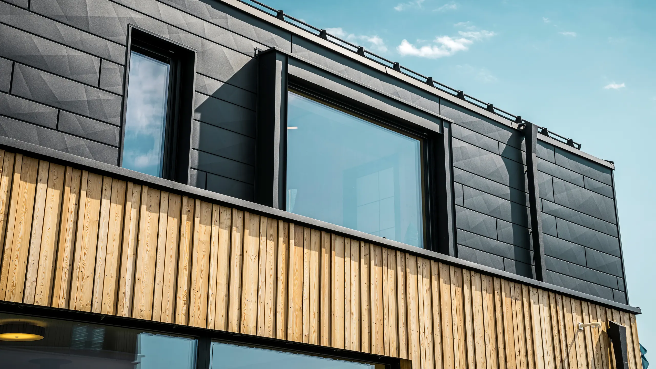 Närbild av övervåningen i modellhuset Autark i prefabricerade huscentret Blaue Lagune, Vösendorf (Österrike). Fasaden består av PREFA Fasadpanel.X aluminiumplattor i P.10 svart, som med sitt speciella utseende med olika kantvinklar verkligen sticker ut. Under dessa finns en stilren träpanel som bildar en harmonisk kontrast till det mörka aluminiumet. Stora fönster släpper in mycket ljus i byggnaden och betonar den moderna designen.