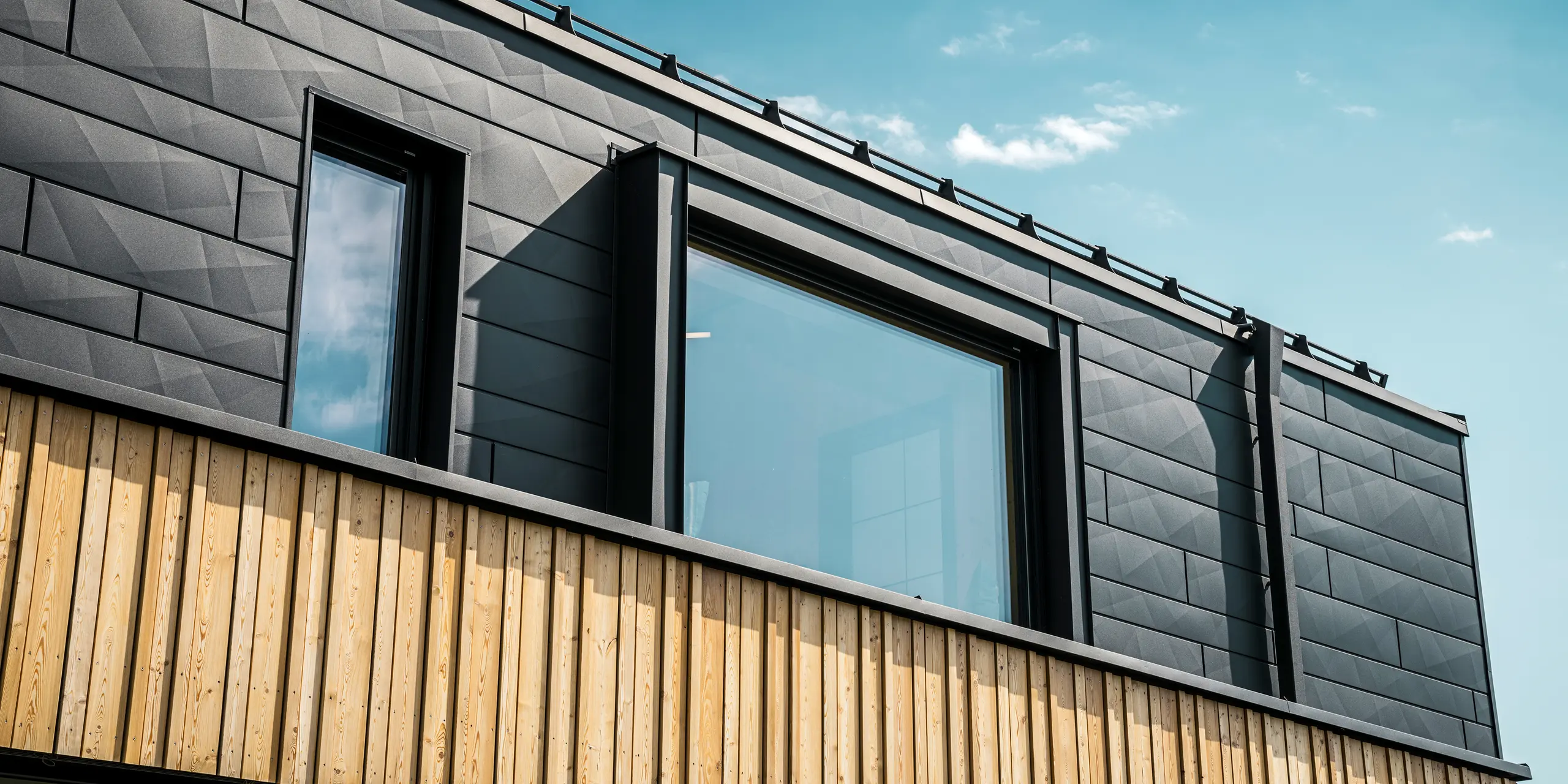 Närbild av övervåningen i modellhuset Autark i prefabricerade huscentret Blaue Lagune, Vösendorf (Österrike). Fasaden består av PREFA Fasadpanel.X aluminiumplattor i P.10 svart, som med sitt speciella utseende med olika kantvinklar verkligen sticker ut. Under dessa finns en stilren träpanel som bildar en harmonisk kontrast till det mörka aluminiumet. Stora fönster släpper in mycket ljus i byggnaden och betonar den moderna designen.
