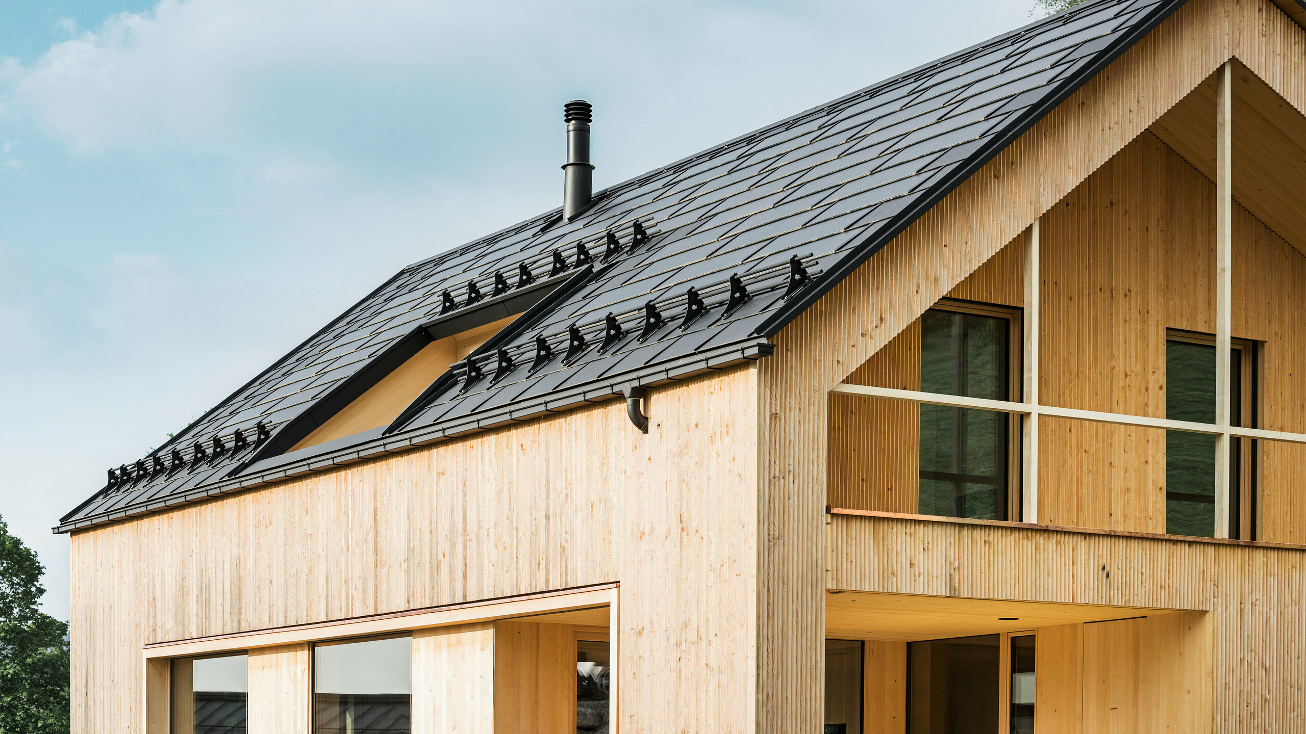 Närbild av gaveln på ett miljövänligt hus i Egg, som kännetecknas av PREFA soltakplattor i elegant svart, som harmoniskt smälter in i mitten av PREFA Takplatta R.16. Takets rena linjer kontrasterar mot den naturliga strukturen på träfasaden, medan solmodulerna är ett tydligt uttryck för hållbart boende. Den diskreta skorstenen smälter in i helhetsbilden, som förenar miljömedvetet design med lantlig estetik. Dessutom har det innovativa ekohuset två balkonger som erbjuder utsikt över den fantastiska bergslandskapet i Egg i Vorarlberg.Närbild av gaveln på ett miljövänligt hus i Egg, som kännetecknas av PREFA soltakplattor i elegant svart, som harmoniskt smälter in i mitten av PREFA Takplatta R.16. Takets rena linjer kontrasterar mot den naturliga strukturen på träfasaden, medan solmodulerna är ett tydligt uttryck för hållbart boende. Den diskreta skorstenen smälter in i helhetsbilden, som förenar miljömedvetet design med lantlig estetik. Dessutom har det innovativa ekohuset två balkonger som erbjuder utsikt över den fantastiska bergslandskapet i Egg i Vorarlberg.