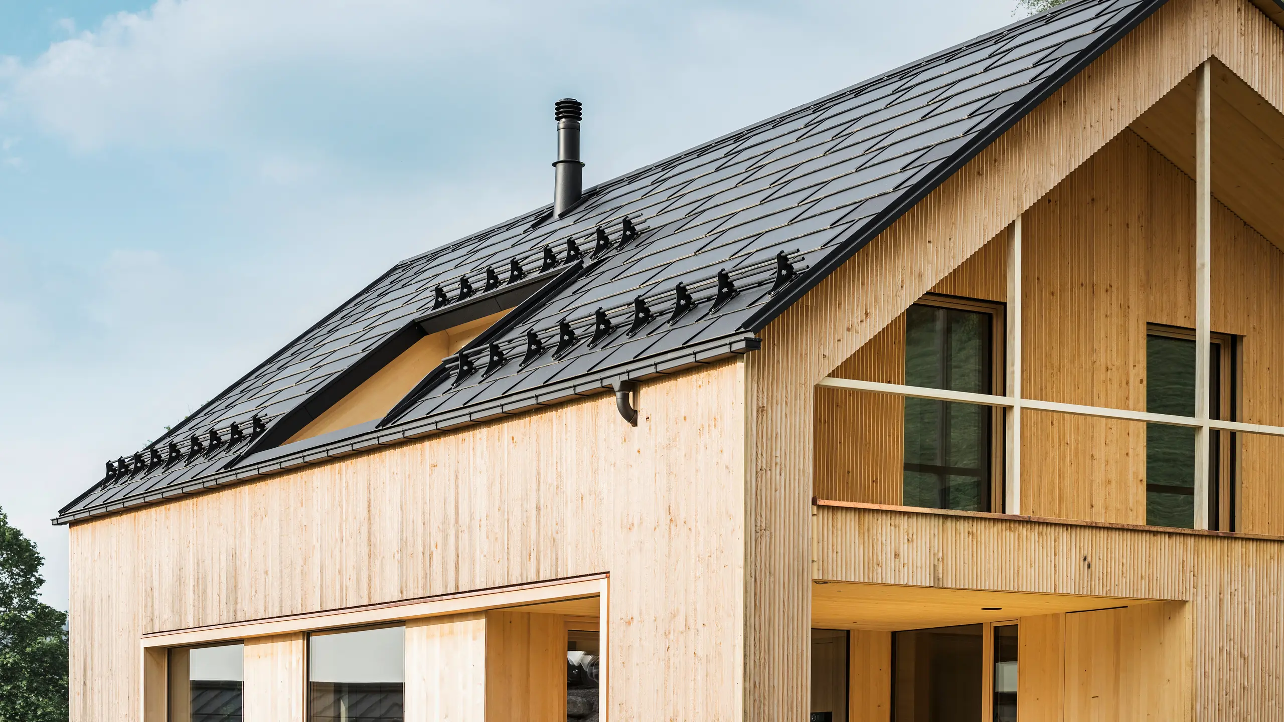 Närbild av gaveln på ett miljövänligt hus i Egg, som kännetecknas av PREFA soltakplattor i elegant svart, som harmoniskt smälter in i mitten av PREFA Takplatta R.16. Takets rena linjer kontrasterar mot den naturliga strukturen på träfasaden, medan solmodulerna är ett tydligt uttryck för hållbart boende. Den diskreta skorstenen smälter in i helhetsbilden, som förenar miljömedvetet design med lantlig estetik. Dessutom har det innovativa ekohuset två balkonger som erbjuder utsikt över den fantastiska bergslandskapet i Egg i Vorarlberg.Närbild av gaveln på ett miljövänligt hus i Egg, som kännetecknas av PREFA soltakplattor i elegant svart, som harmoniskt smälter in i mitten av PREFA Takplatta R.16. Takets rena linjer kontrasterar mot den naturliga strukturen på träfasaden, medan solmodulerna är ett tydligt uttryck för hållbart boende. Den diskreta skorstenen smälter in i helhetsbilden, som förenar miljömedvetet design med lantlig estetik. Dessutom har det innovativa ekohuset två balkonger som erbjuder utsikt över den fantastiska bergslandskapet i Egg i Vorarlberg.