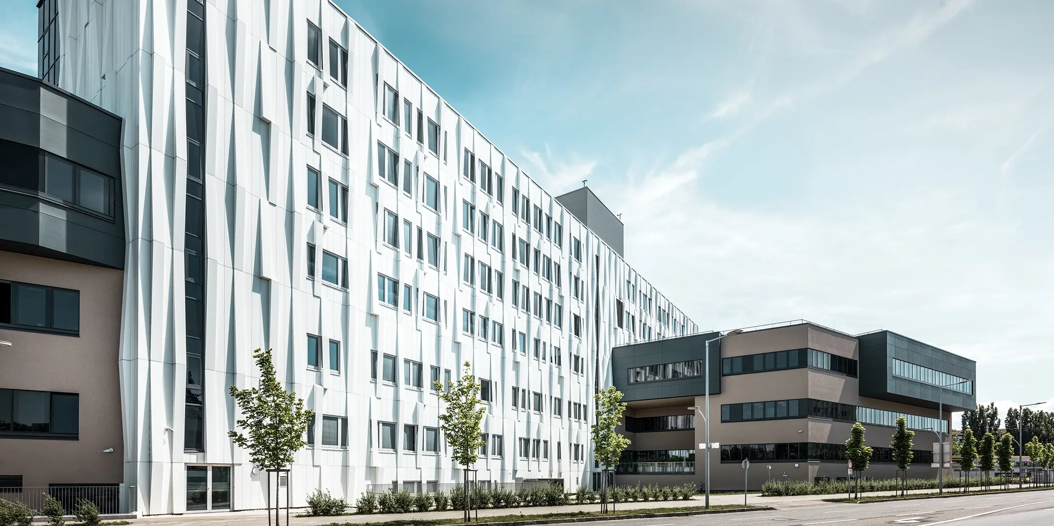 Sidovy av universitetssjukhuset i St. Pölten, Niederösterreich. Den robusta aluminiumfasaden gör byggnaden till ett blickfång i staden. I förgrunden syns en gata med några träd.