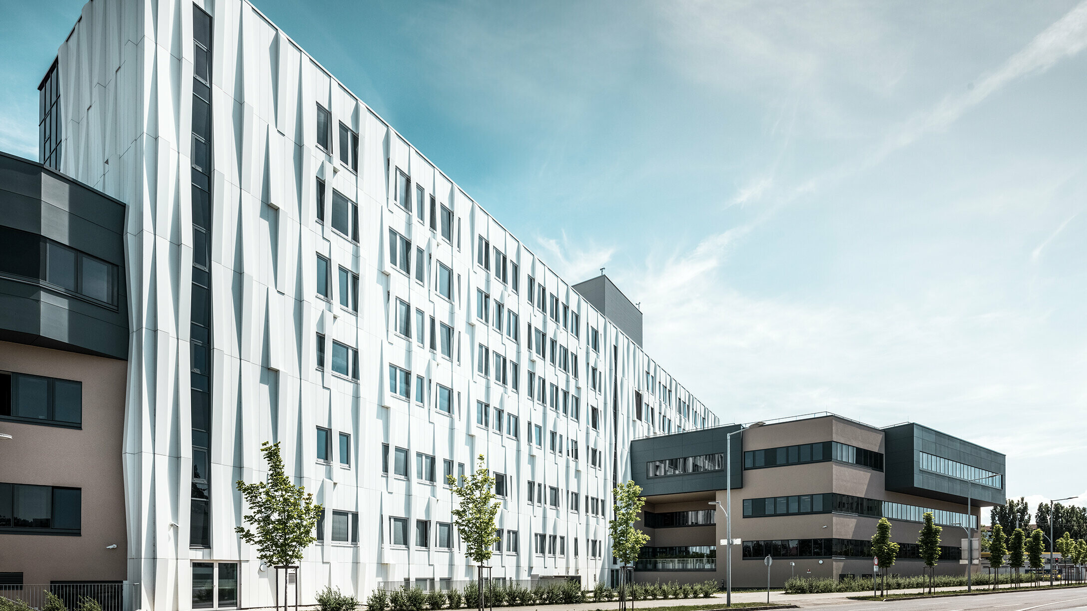 Sidovy av universitetssjukhuset i St. Pölten, Niederösterreich. Den robusta aluminiumfasaden gör byggnaden till ett blickfång i staden. I förgrunden syns en gata med några träd.