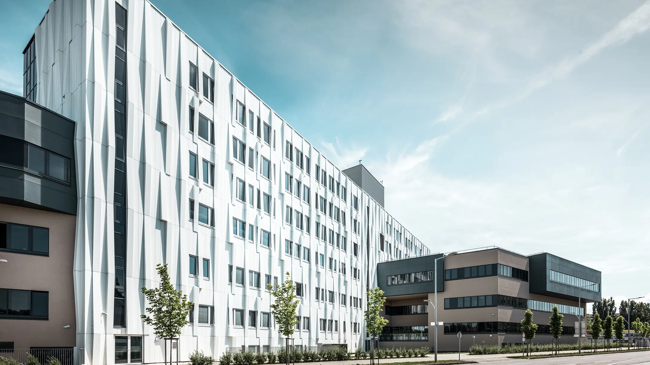 Sidovy av universitetssjukhuset i St. Pölten, Niederösterreich. Den robusta aluminiumfasaden gör byggnaden till ett blickfång i staden. I förgrunden syns en gata med några träd.
