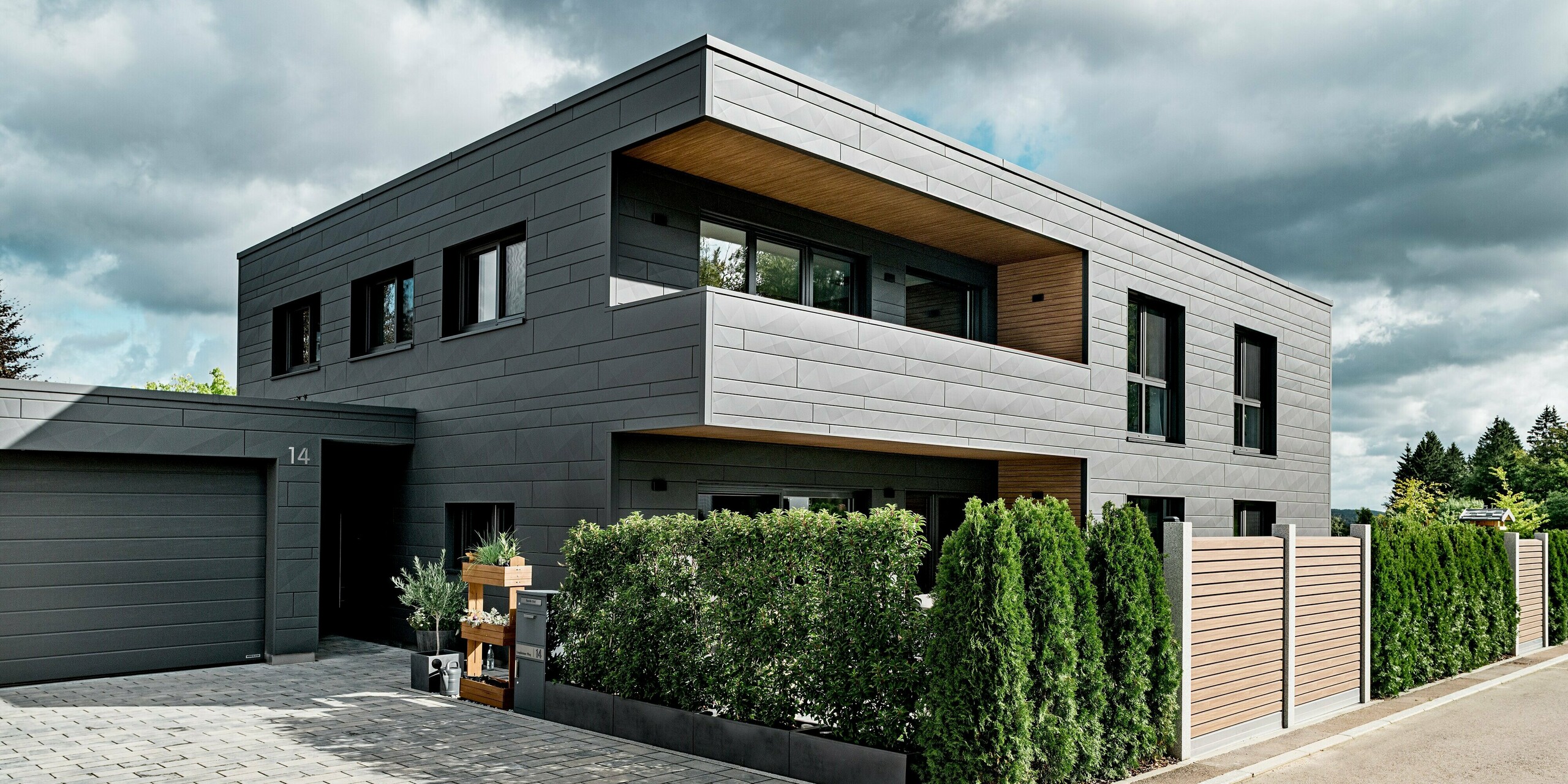 Ansicht der Zufahrt eines modernen Einfamilienhauses in Heidenheim mit vollständig verkleideter PREFA Siding.X Aluminiumfassade in P.10 Dunkelgrau. Die klar strukturierten Paneele mit unterschiedlichen Breiten und markanten Kantungen erzeugen ein dynamisches Fassadenbild. Eine dichte Hecke und ein Holzzaun setzen natürliche und warme Kontraste zur präzisen Metalloberfläche. Die überdachte Eingangszone sowie die große Garage fügen sich harmonisch ins Gesamtbild ein. Die langlebige PREFA Aluminiumoberfläche bietet dauerhaften Schutz und moderne, hochwertige Architektur.