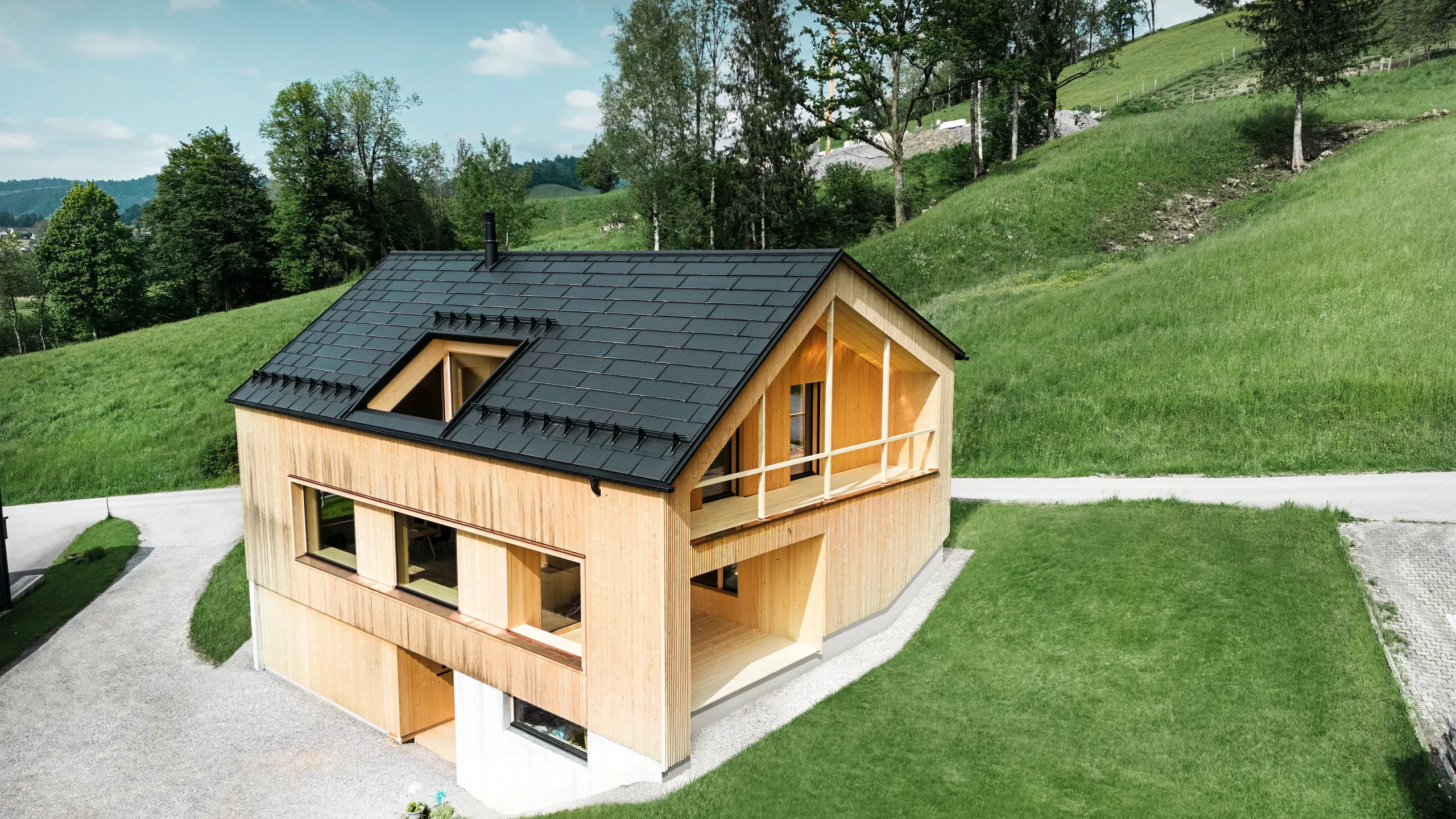 Bilden visar ett pittoreskt ekohus i Egg, inbäddat i de mjuka gröna kullarna, med ett hållbart PREFA-soldak. De svarta, högkvalitativa PREFA-takplattorna R.16 och PREFA-solar dakplattorna smälter harmoniskt in i den lantliga landskapet och framhäver omgivningens naturliga skönhet. Solpanelerna är inte bara ett engagemang för förnybar energi, utan också ett designelement som stilfullt kompletterar den moderna träfasaden och byggnadens rena linjer. Detta hus är ett föredöme för miljömedvetet byggande och en inspiration för framtidsinriktad arkitektur.