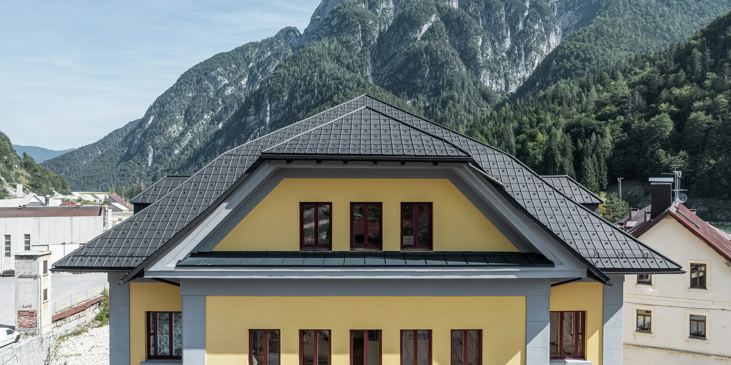 Nahaufnahme der Dacheindeckung des Museo Cave del Predil in Italien. Das gelbe Museumsgebäude wurde mit PREFA Dachplatten in P.10 Anthrazit eingedeckt. Die robuste Dachlösung aus Aluminium präsentiert sich harmonisch auf dem historischen Gebäude mit gelber Fassade. Im Hintergrund die beeindruckende Alpenlandschaft mit bewaldeten Berghängen, die eine extreme Witterungsbeständigkeit der PREFA Produkte erfordert.