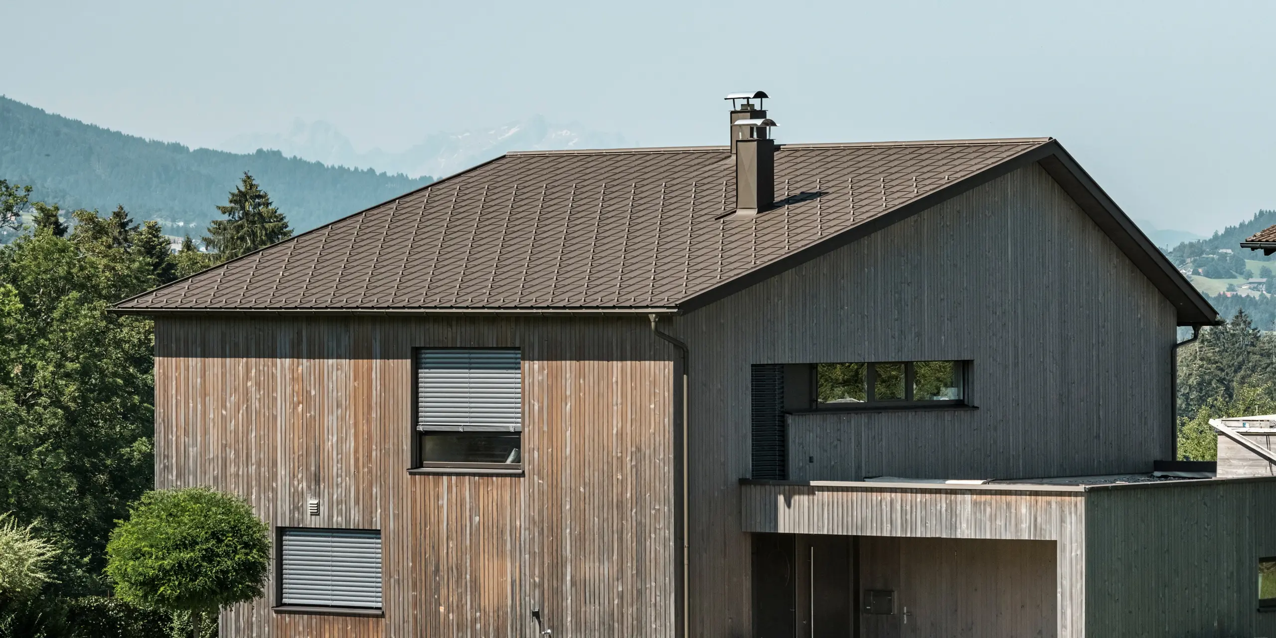 Sidovy av ett modernt enfamiljshus i Krumbach med en kombination av en naturlig träfasad och ett hållbart PREFA-tak i aluminium i P.10 brun. Det harmoniska färgschemat visar den eleganta integrationen av modern design och traditionella material, med PREFA takplattor som ger en estetisk och funktionell takdesign.