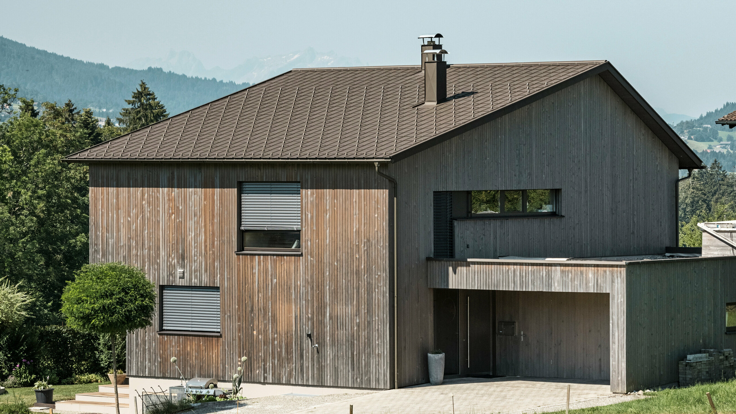 Sidovy av ett modernt enfamiljshus i Krumbach med en kombination av en naturlig träfasad och ett hållbart PREFA-tak i aluminium i P.10 brun. Det harmoniska färgschemat visar den eleganta integrationen av modern design och traditionella material, med PREFA takplattor som ger en estetisk och funktionell takdesign.