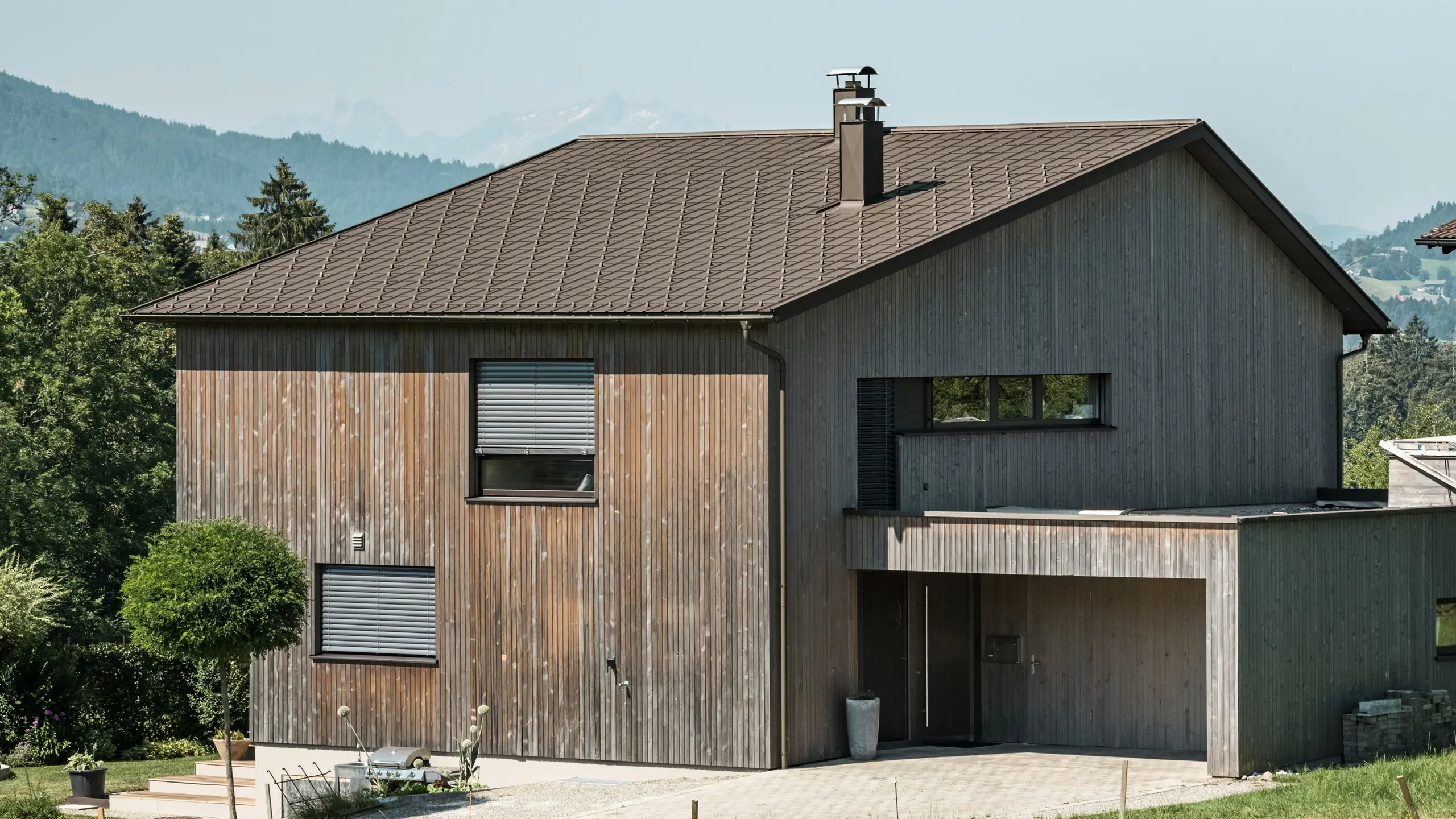 Sidovy av ett modernt enfamiljshus i Krumbach med en kombination av en naturlig träfasad och ett hållbart PREFA-tak i aluminium i P.10 brun. Det harmoniska färgschemat visar den eleganta integrationen av modern design och traditionella material, med PREFA takplattor som ger en estetisk och funktionell takdesign.