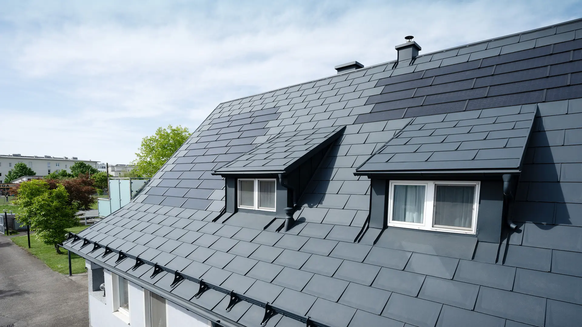 Närbild av ett tak täckt med stora PREFA-solar takplattor och PREFA-takplattor av typen R.16 i färgen P.10 antracit. Två takkupor syns. Solar takplattorna är jämnt fördelade över taket och skapar en modern, hållbar lösning.