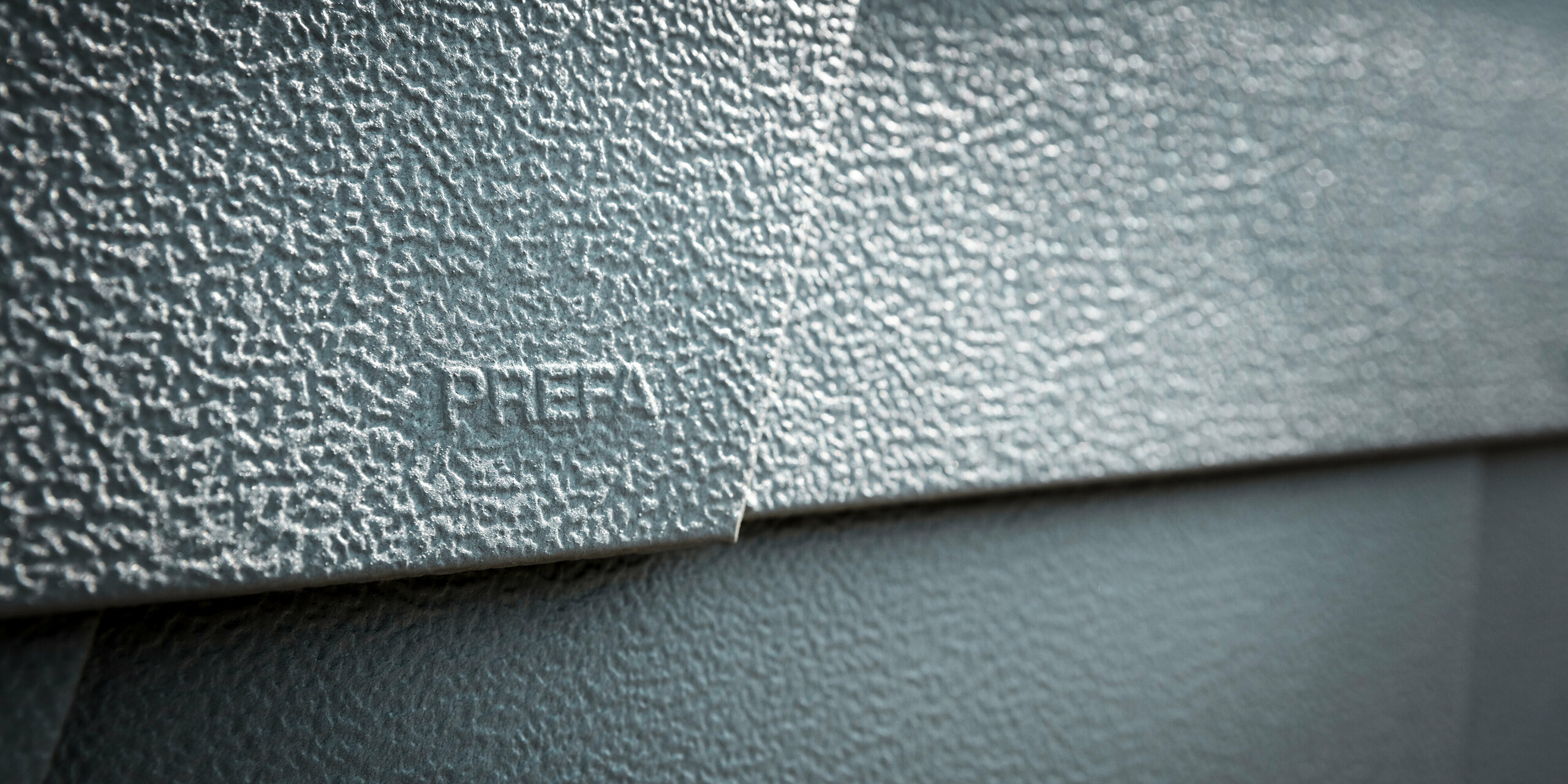 Detailaufnahme einer PREFA Dachschindel in P.10 Hellgrau, die mit ihrer strukturierten Oberfläche und dem eingestanzten Wort „PREFA“ Qualität und Markenbewusstsein demonstriert. Diese Aufnahme zeigt die robuste Beschaffenheit und die anspruchsvolle Textur der Aluminiumschindel, welche die Langlebigkeit und Witterungsbeständigkeit von PREFA Produkten widerspiegelt und für zeitgenössische sowie funktionale Dach- und Fassadengestaltung steht.