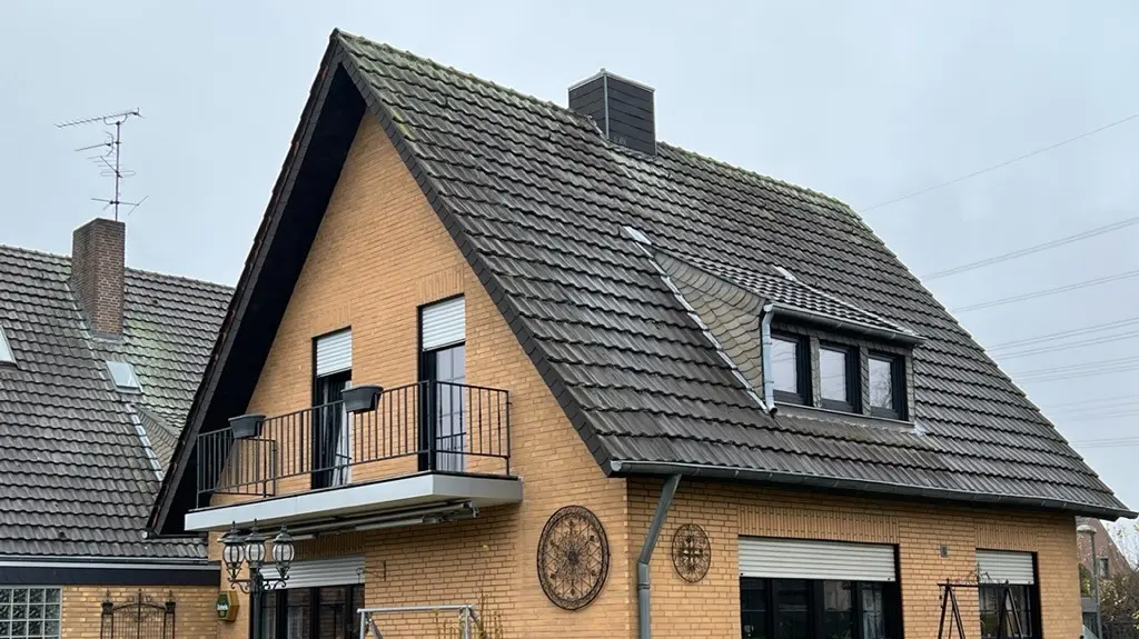 Enfamiljshus i gul klinker med brant sadeltak och mörk takbeläggning före takrenoveringen; på trädgårdssidan med balkong, takkupol, terrass med sittmöbler och gräsmatta i förgrunden.
