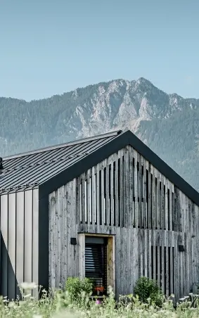 Moderner, funktionaler Wohnbau mit einer Kombination aus Holzfassade und PREFA Band- und Blechmaterial für das Dach. Die klare Architektur wird durch die elegante, metallische Dach- und Fassadenverkleidung unterstrichen, die ideal für anspruchsvolle Projekte ist. Das Bild veranschaulicht die vielseitigen Anwendungsmöglichkeiten der PREFA Produkte im Bereich Bänder und Bleche.
