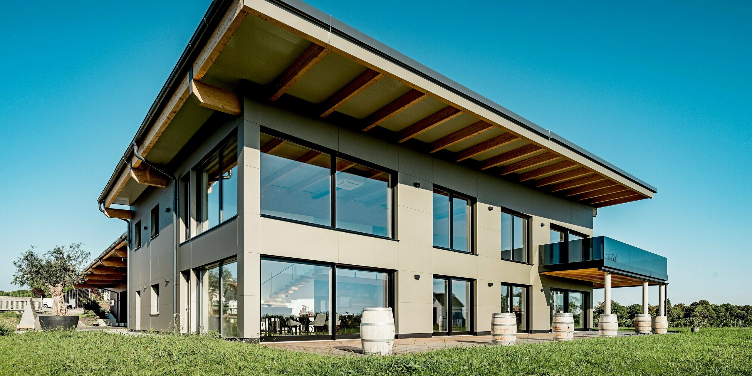 Diese Weitwinkelaufnahme zeigt das moderne Weingut Nigl in Perchtoldsdorf mit seiner markanten Aluminiumfassade in der Farbe P.10 Prefabronze. Zu sehen ist die Rückseite des Gebäudes mit klar gegliederter Fassadenstruktur, bodentiefen Glasflächen, einem weit auskragenden Pultdach mit sichtbaren Holzbalken sowie einem eleganten Balkon mit Glasbrüstung. Die Aluminium Verbundplatten verleihen dem Baukörper eine edle, ruhige Optik und setzen einen spannenden Kontrast zur umliegenden Natur. Das Lichtspiel auf der metallischen Oberfläche betont die hochwertige Verarbeitung der PREFA Produkte.