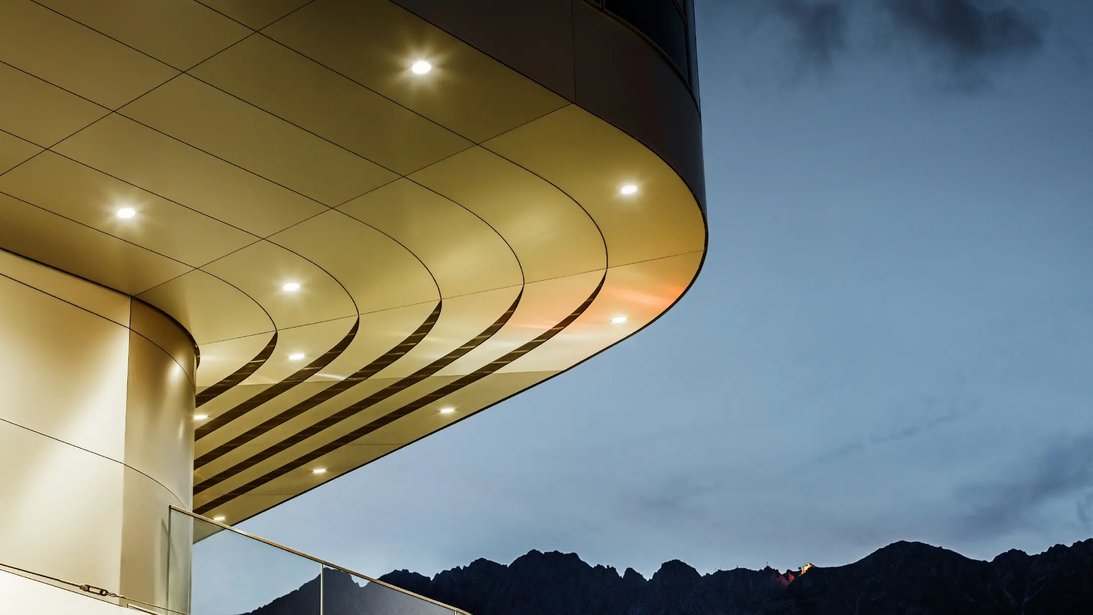 [Translate to Svenska:] Das Bild zeigt eine beeindruckende architektonische Detailsansicht einer PREFA PEMA Verbundplatte aus gebürstetem Aluminium. Die Platte ist sanft gewölbt und verläuft in eleganten, fließenden Linien, die durch gezielte Beleuchtung am Abend besonders zur Geltung kommen. Die klare Struktur und der edle Aluminium-Look der Platte ergänzen das moderne Design der Fassade und bieten eine elegante, zeitgenössische Lösung für hochwertige Architektur.
