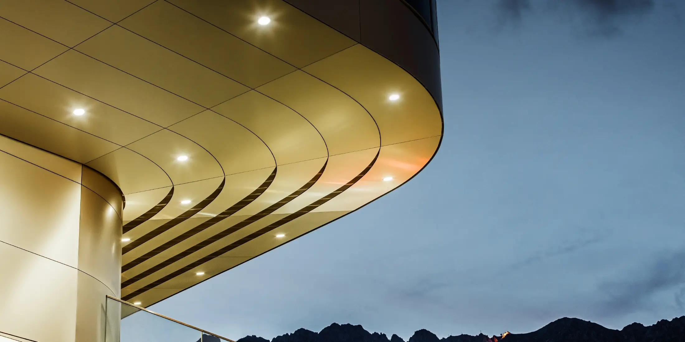 [Translate to Svenska:] Das Bild zeigt eine beeindruckende architektonische Detailsansicht einer PREFA PEMA Verbundplatte aus gebürstetem Aluminium. Die Platte ist sanft gewölbt und verläuft in eleganten, fließenden Linien, die durch gezielte Beleuchtung am Abend besonders zur Geltung kommen. Die klare Struktur und der edle Aluminium-Look der Platte ergänzen das moderne Design der Fassade und bieten eine elegante, zeitgenössische Lösung für hochwertige Architektur.