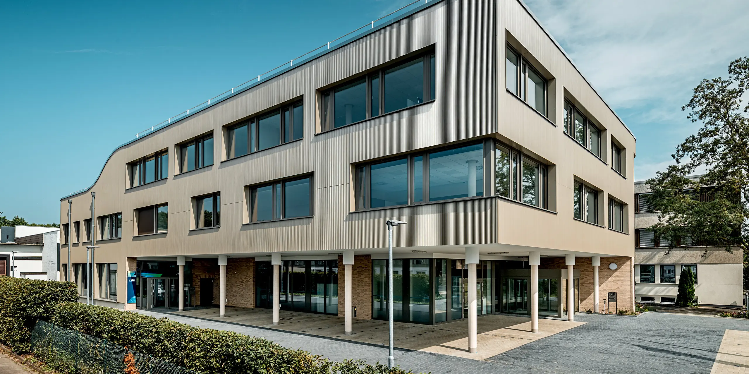 Das Bild zeigt den Eingangsbereich des modernen Festo-Gebäudes in Langenhagen, das durch eine markante, geschwungene Fassade besticht. Die Fassade ist mit PREFA Strangpressprofilen in Zackenform verkleidet, die eine interessante Textur und Tiefe erzeugen. Die klaren Fensterflächen und die glatte, elegante Struktur der Aluminium Verbundplatten sorgen für eine harmonische Kombination aus Funktionalität und Design. Besonders hervorzuheben ist die sanft abgerundete, obere Kante des Gebäudes, die dem gesamten Bauwerk eine moderne, fließende Ausstrahlung verleiht. 