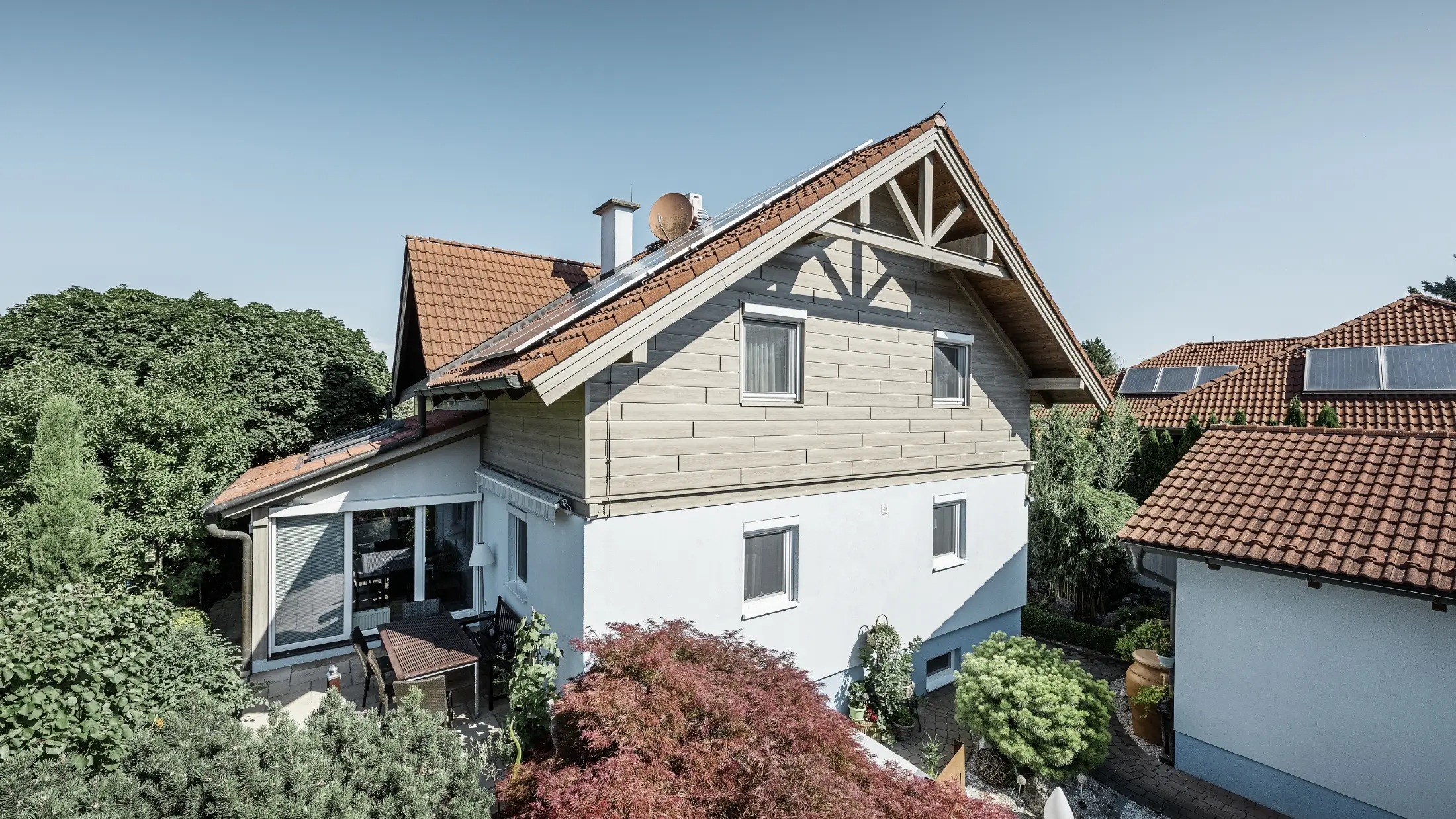 Enfamiljshus med sadeltak och rött tak, kombinerat med en modern fasaddesign. Den övre gaveln är klädd med beige-grå aluminiumfasadpaneler från PREFA, medan bottenvåningen har en vit putsad fasad. På taket finns solcellspaneler. Huset ligger i en grön trädgård med terrass, trädgårdsmöbler och frodig växtlighet, omgivet av andra bostadshus med liknande takarkitektur.