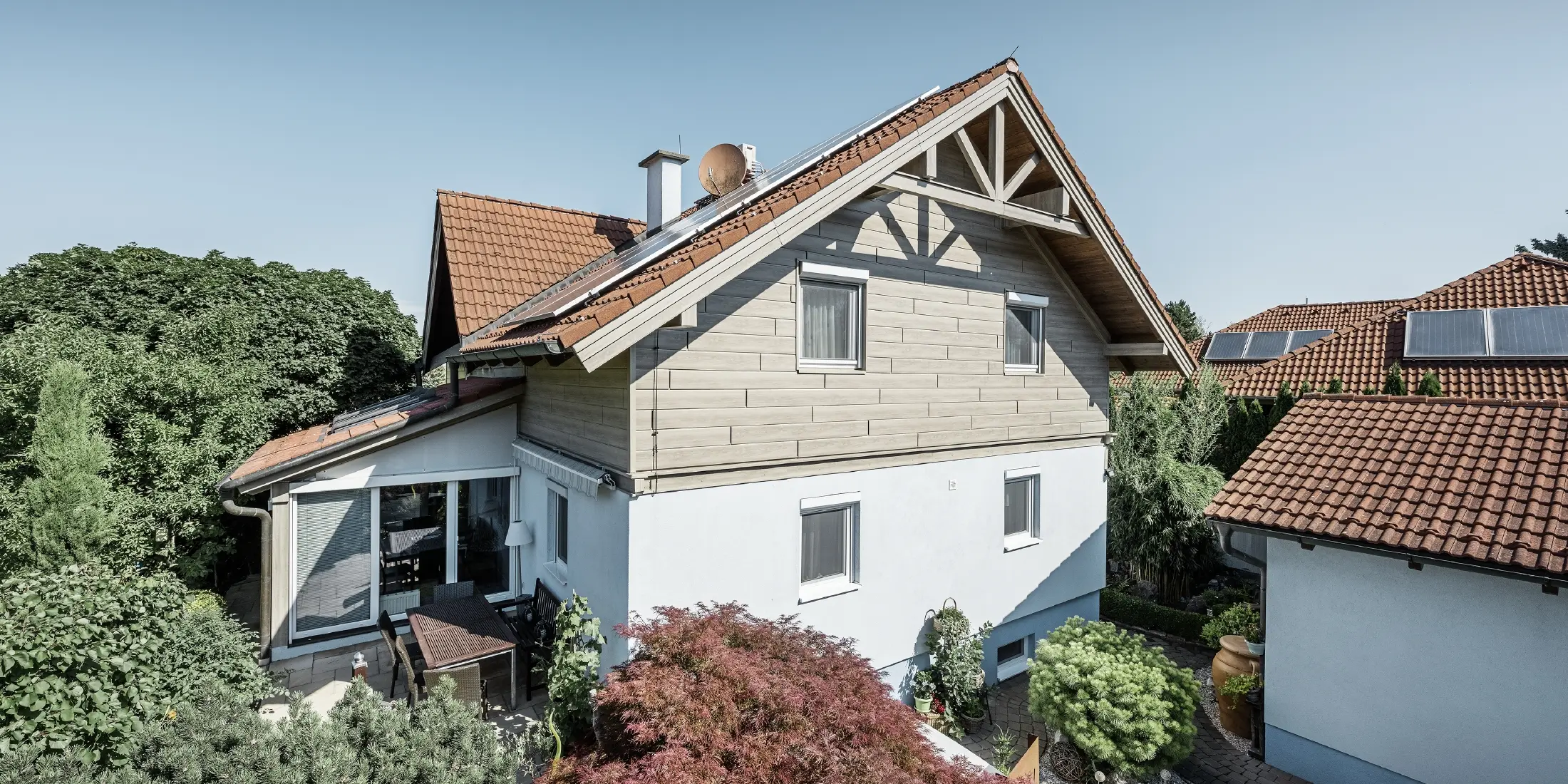 Enfamiljshus med sadeltak och rött tak, kombinerat med en modern fasaddesign. Den övre gaveln är klädd med beige-grå aluminiumfasadpaneler från PREFA, medan bottenvåningen har en vit putsad fasad. På taket finns solcellspaneler. Huset ligger i en grön trädgård med terrass, trädgårdsmöbler och frodig växtlighet, omgivet av andra bostadshus med liknande takarkitektur.