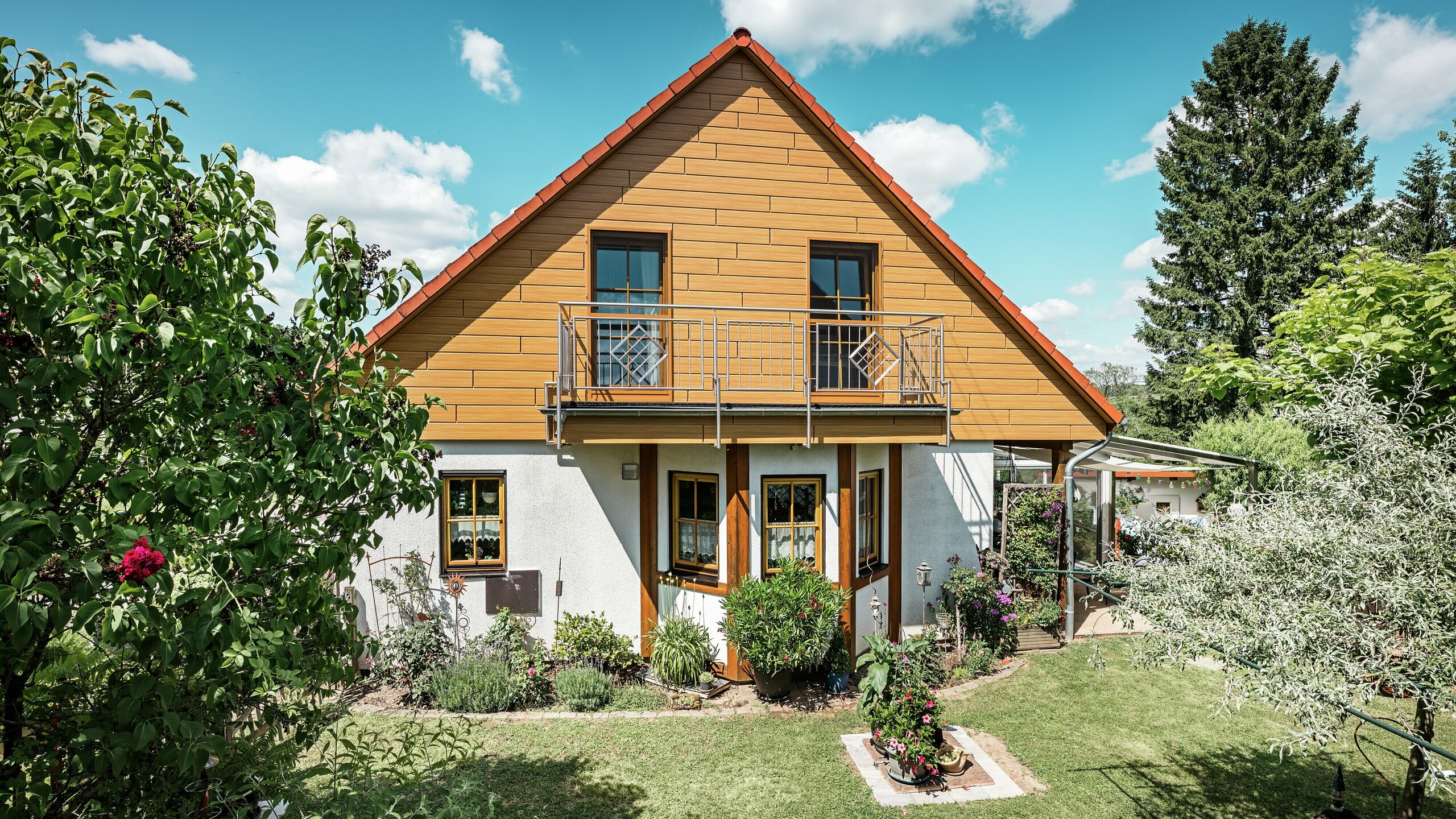 På denna bild lyser den renoverade gaveln på en villa i Blieskastel i en varm, naturlig ek natur-färg tack vare PREFA Siding. Den högkvalitativa aluminiumfasaden med realistisk trälook sätter en stark visuell prägel och harmonierar imponerande med det röda tegeltaket och den frodiga trädgården. Omgiven av blommor, buskar och frodig grönska ger huset ett inbjudande och välskött intryck. PREFA-ytan kombinerar ett naturligt utseende med funktionella fördelar som hög väderbeständighet, stabilitet och praktiskt taget underhållsfri hållbarhet – en estetisk höjdpunkt med hållbart mervärde.