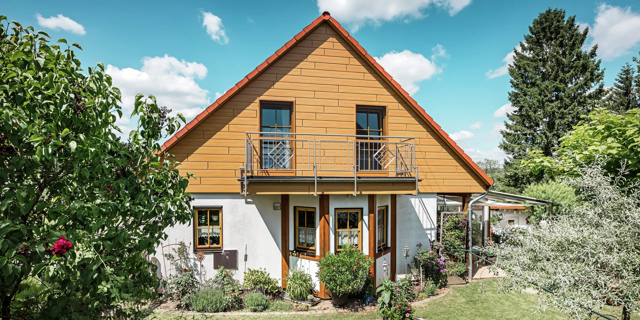 På denna bild lyser den renoverade gaveln på en villa i Blieskastel i en varm, naturlig ek natur-färg tack vare PREFA Siding. Den högkvalitativa aluminiumfasaden med realistisk trälook sätter en stark visuell prägel och harmonierar imponerande med det röda tegeltaket och den frodiga trädgården. Omgiven av blommor, buskar och frodig grönska ger huset ett inbjudande och välskött intryck. PREFA-ytan kombinerar ett naturligt utseende med funktionella fördelar som hög väderbeständighet, stabilitet och praktiskt taget underhållsfri hållbarhet – en estetisk höjdpunkt med hållbart mervärde.