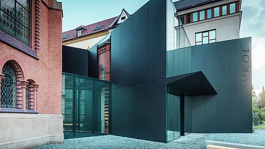 Das Bild zeigt die moderne Erweiterung einer Bibliothek, die sich elegant an die historische Rote Kirche in Olmütz anschließt. Der markante Neubau beeindruckt mit einer Fassade aus schwarzen PREFABOND Aluminium-Verbundplatten, die in klaren, geometrischen Formen gestaltet ist und mit präziser Detailarbeit überzeugt. Die Glaswände des Gebäudes bieten einen transparenten Blick in das Innere, das sich harmonisch in die historische Umgebung einfügt. Das Zusammenspiel von modernen Materialien und klassischen Architekturelementen schafft eine spannende visuelle Verbindung zwischen Alt und Neu. Der innovative Einsatz der PREFABOND Platten sorgt nicht nur für ein modernes Erscheinungsbild, sondern auch für Langlebigkeit und Widerstandsfähigkeit, die das Gebäude für die Zukunft wappnen.