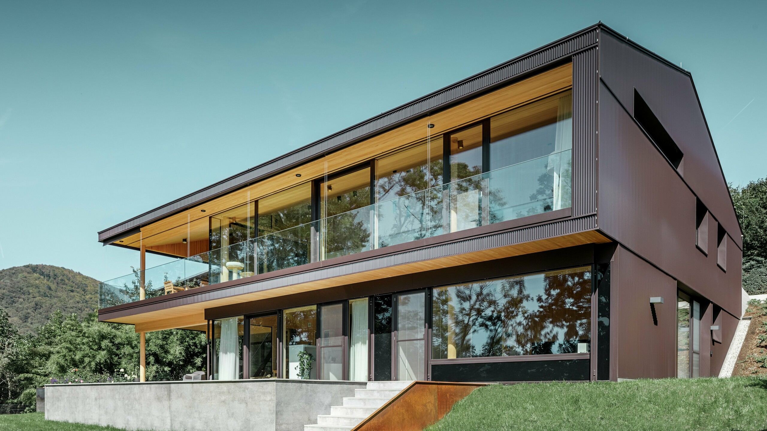 Modernt fristående hus med stora fönster på trädgårdssidan, fasaden kläddes med PREFA sicksackprofil i mörkbrunt.