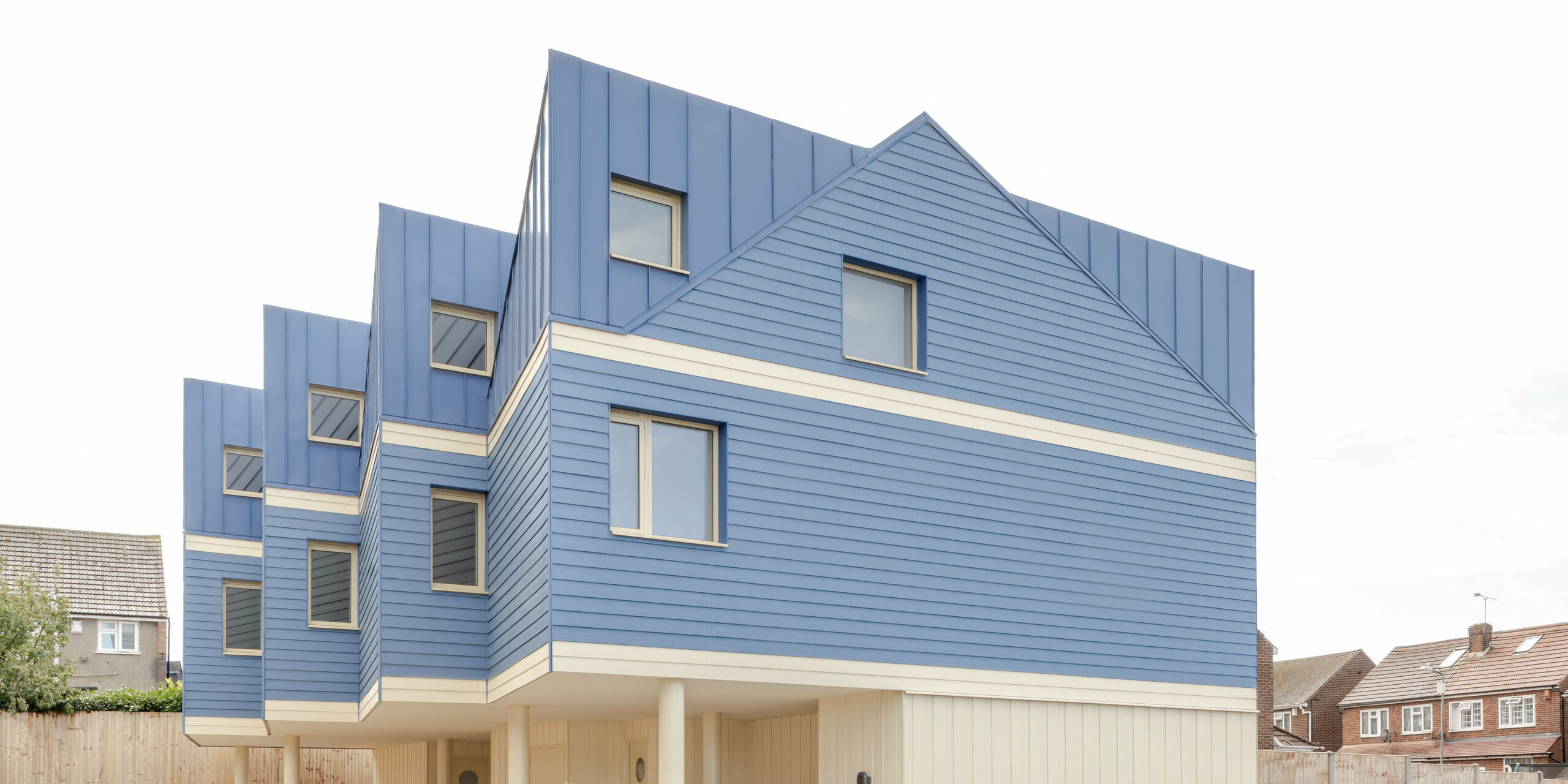 Das Bild zeigt die markante Vorderseite der Goldsmith‑Mews‑Reihenhausanlage mit ihrer auffälligen FALZONAL Verkleidung in Taubenblau. Die präzisen, vertikalen Falzbahnen im Dach- und Obergeschoss gehen harmonisch in die horizontalen Aluminiumprofile über und erzeugen ein lebendiges, rhythmisches Fassadenbild. Die hellen unteren Bereiche und die aufgeständerte Konstruktion verleihen dem Gebäude Leichtigkeit. Das langlebige, farbstabile PREFA Aluminium bietet zuverlässigen Witterungsschutz, hohe Formbeständigkeit und ermöglicht eine moderne, klare Architektur mit starker Wiedererkennung.