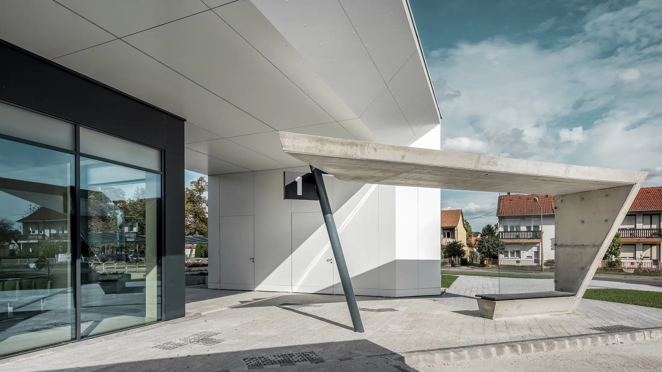 Vstupná časť autobusovej stanice Lenti; spodná strana strechy je obložená hliníkovým kompozitným panelom PREFABOND od spoločnosti PREFA v čisté bielej farbe, fasádne prvky pri vchodových dverách a výkladoch sú obložené v P.10 antracitovej farbe.