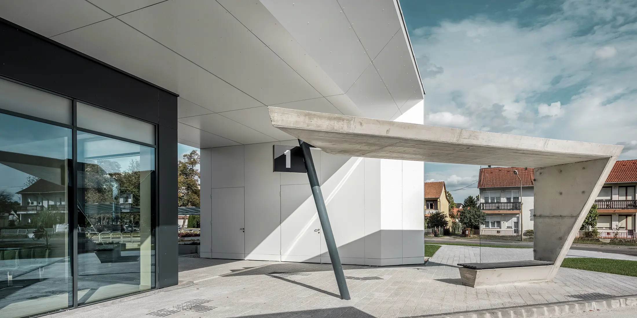 Vstupná časť autobusovej stanice Lenti; spodná strana strechy je obložená hliníkovým kompozitným panelom PREFABOND od spoločnosti PREFA v čisté bielej farbe, fasádne prvky pri vchodových dverách a výkladoch sú obložené v P.10 antracitovej farbe.