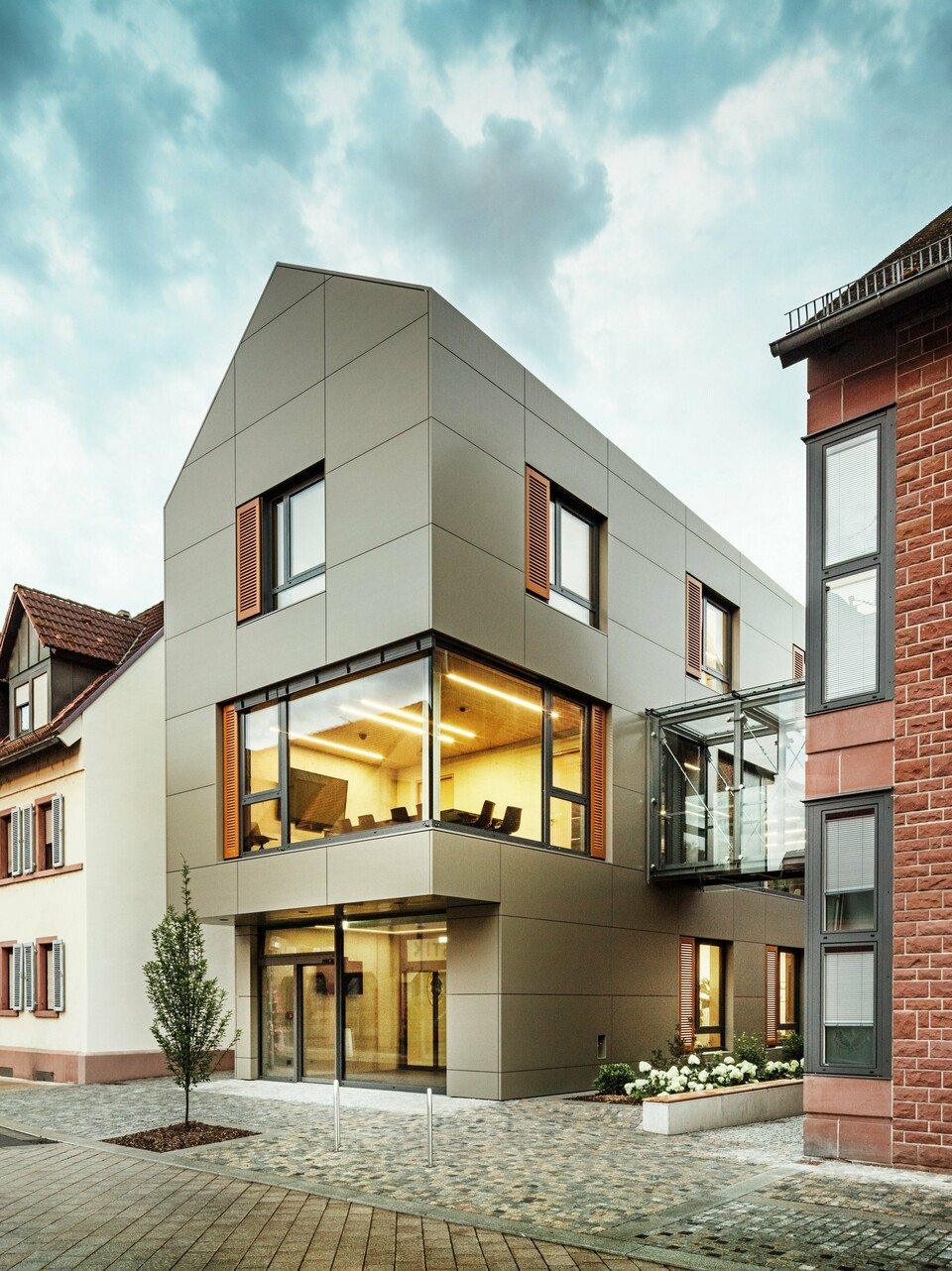 Slika prikazuje modernu gradsku vijećnicu u Alzenauu, obloženu aluminijskim kompozitnim pločama PREFA u toploj brončanoj nijansi. Fasadu prekidaju velike staklene površine koje otkrivaju unutrašnjost obasjanu toplim svjetlom. Stakleni spojni hodnik na desnoj strani slike povezuje zgradu s obližnjom tradicionalnom zgradom od opeke, stvarajući zanimljiv kontrast između moderne i povijesne arhitekture.