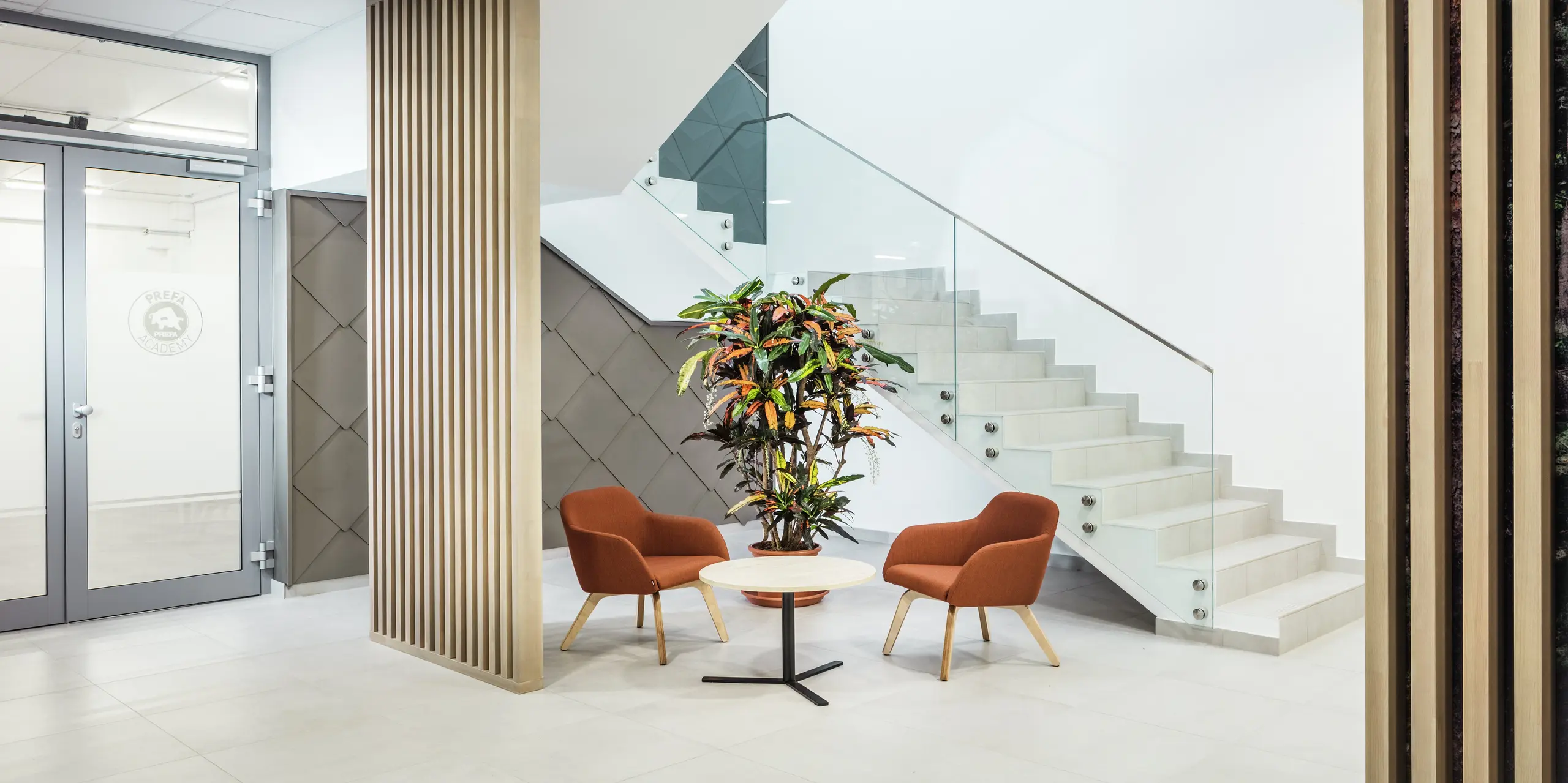 Das stilvolle Interieur des neuen PREFA Standortes in Törökbálint (Ungarn) überzeugt durch eine moderne und einladende Gestaltung. Neben dem Eingang zu den praktischen Schulungsräumen der PREFA Academy wurde die Wandverkleidung mit PREFA Wandrauten 44 × 44 in Bronze akzentuiert. Der Eyecatcher zieht sich von der Außenfassade bis ins Innere und verbindet dabei Design mit Funktionalität. Auch im Innenbereich fügt sich die hochwertige Aluminiumoberfläche harmonisch in das Gesamtkonzept ein. Neben hellen Holzelementen wurden einladende Sitzmöbel und Grünpflanzen platziert, um den Zugang zu den Büros über das Treppenhaus für die Mitarbeiterinnen und Mitarbeiter angenehmer zu gestalten.