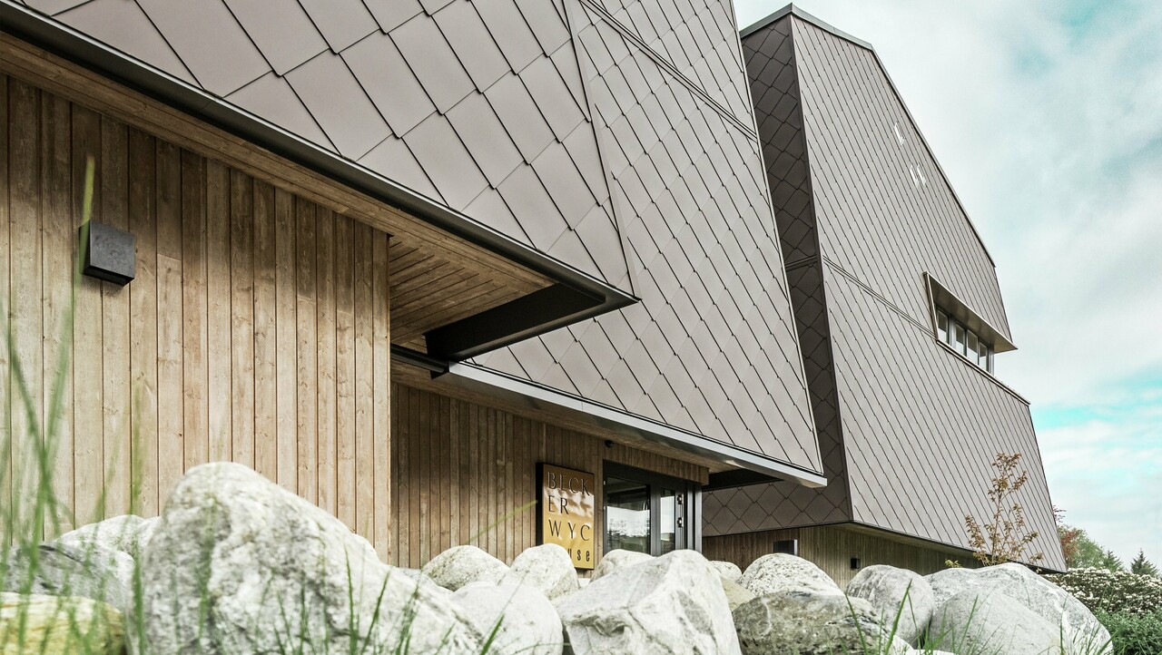 Sidovy av ett hotell, bottenvåningens fasad är vertikalt klädd med mörkfärgat trä. Ovanför detta finns ett fasadutsprång, härifrån är fasaden klädd med aluminiumpaneler i valnötsbrunt.