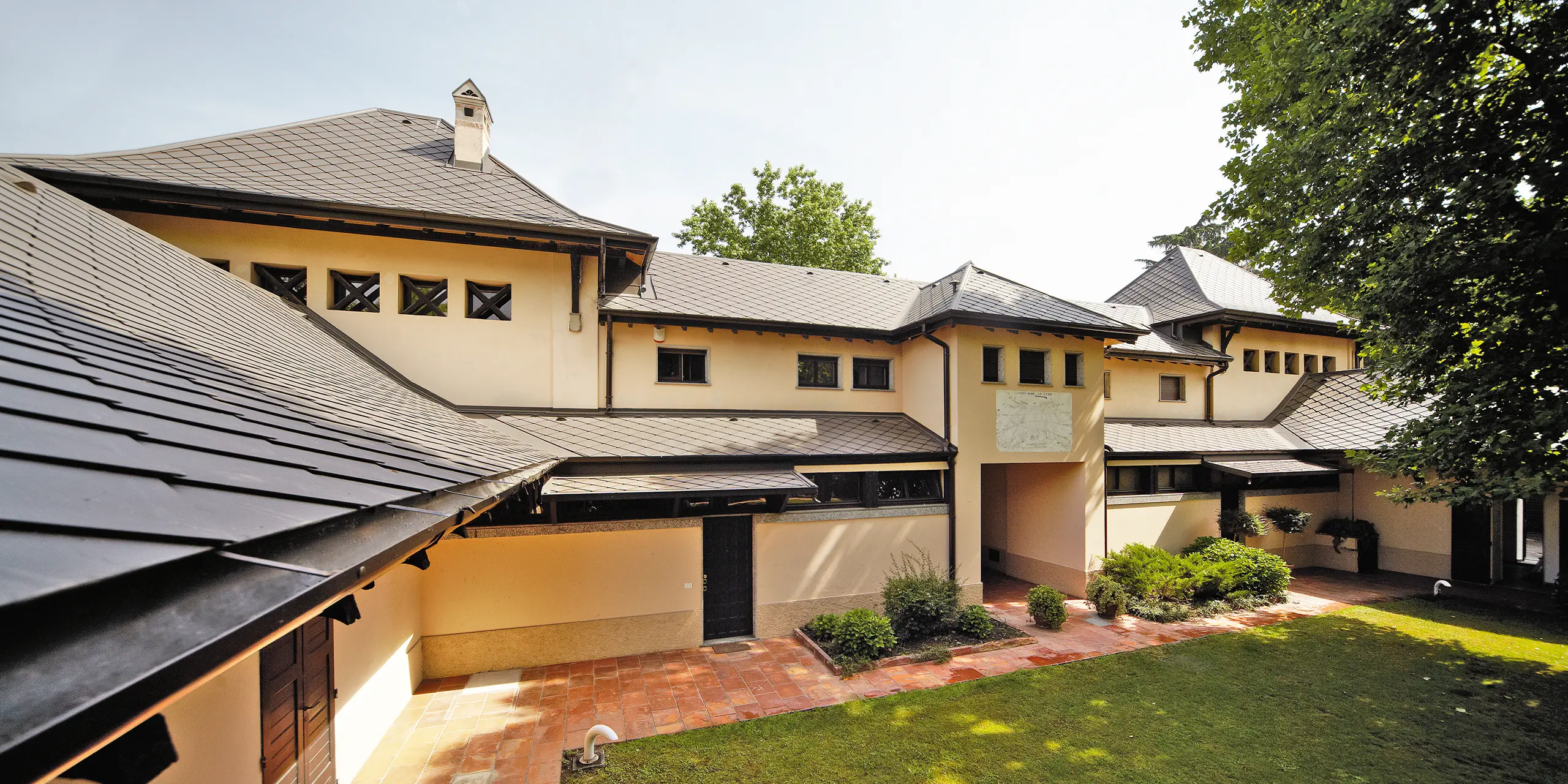 Italiensk villa med rombtak i P.10 brun från PREFA med beige fasadfärg och skuggig innergård