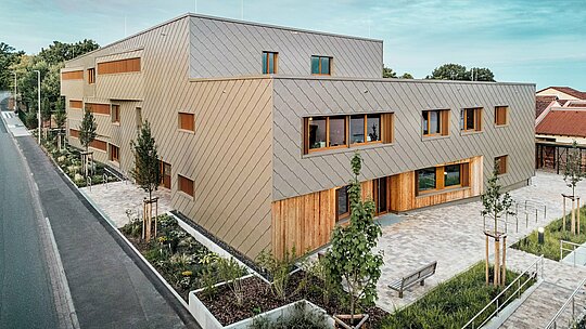 Die Abendstimmung taucht die Kindertagesstätte in Alzenau in ein sanftes Licht und betont die hochwertige Fassade aus bronzefarbenen PREFA Wandrauten 44 × 44. Die Kombination aus strukturierter Aluminiumfassade und vertikalen Holzflächen schafft ein lebendiges Wechselspiel aus Material, Farbe und Textur. Großzügige Fenster sorgen für helle Innenräume, während die umliegenden Grünanlagen und der gepflasterte Vorplatz zum Verweilen einladen. Das Bild zeigt eindrucksvoll, wie zeitgemäße Architektur, Funktionalität und Nachhaltigkeit mit PREFA Produkten harmonisch umgesetzt werden – mitten im städtischen Kontext von Alzenau.