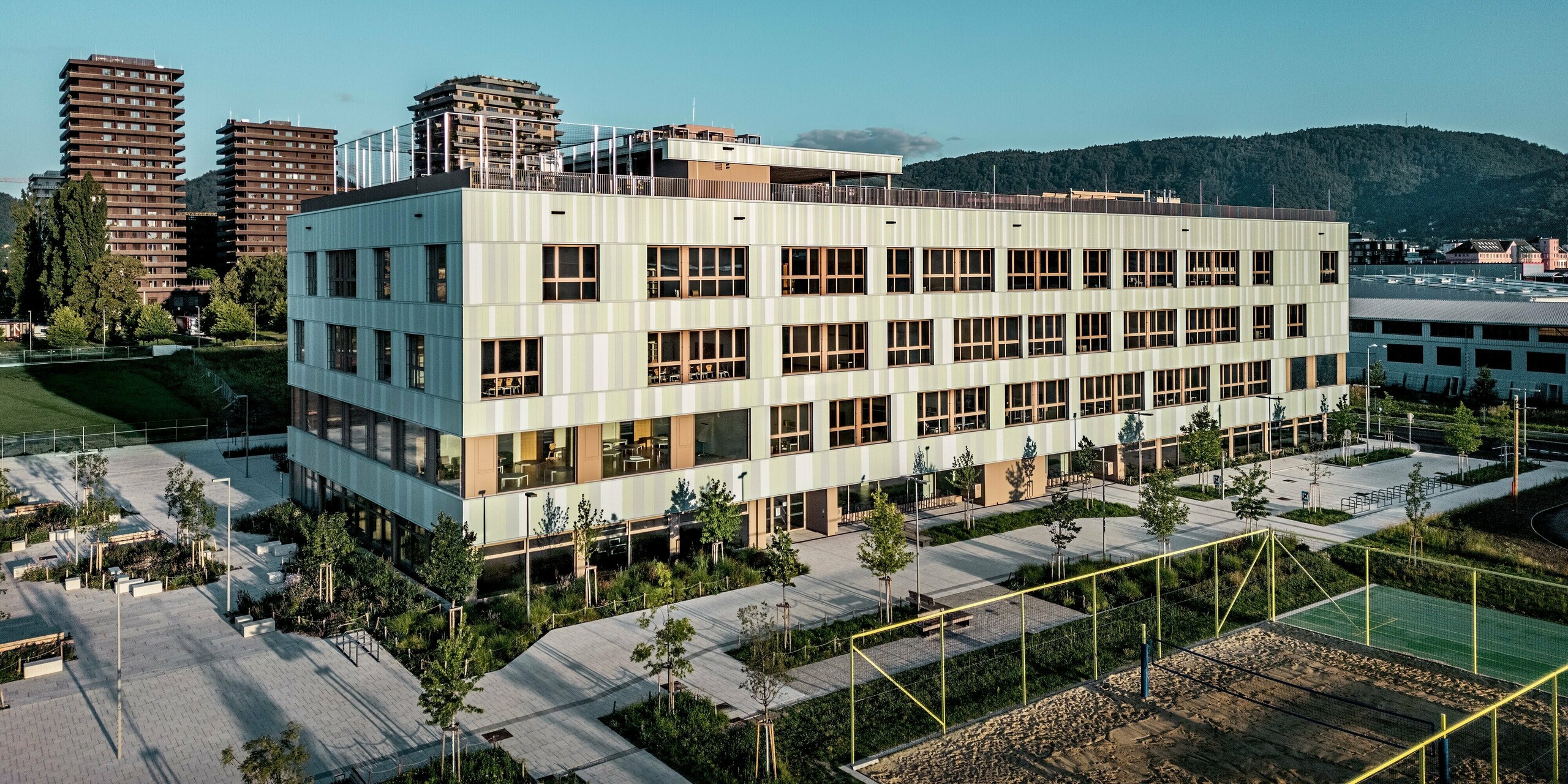 Das Bild zeigt die AHS Reininghaus in Graz aus der Vogelperspektive. Die PREFA Sidings in Sonderfarben, die das Gebäude schmücken, bestehen aus sanften Grüntönen und Weiß und sind in vertikalen Streifen angeordnet. Diese Farbwahl sorgt für eine frische, moderne Ausstrahlung und lässt das Gebäude lebendig und zugleich elegant wirken. Die braunen Fensterrahmen bilden einen schönen Kontrast zur helleren Fassadenfarbe und tragen zur stilvollen Gesamtwirkung bei. Im Vordergrund sind großzügige Außenbereiche mit gepflegten Grünflächen, Sitzbänken und einem Sportplatz zu sehen. Ein Volleyballplatz und ein gepflegtes Gehwegnetz fördern eine angenehme und funktionale Nutzung des Außengeländes. Die Umgebung wird durch moderne, höher gelegene Gebäude ergänzt, die dem architektonischen Kontext der AHS Reininghaus einen urbanen Charakter verleihen. Die Architektur der Schule fügt sich perfekt in das urbane Umfeld ein und ist ein markantes Beispiel für zeitgemäße Bildungseinrichtungsarchitektur.