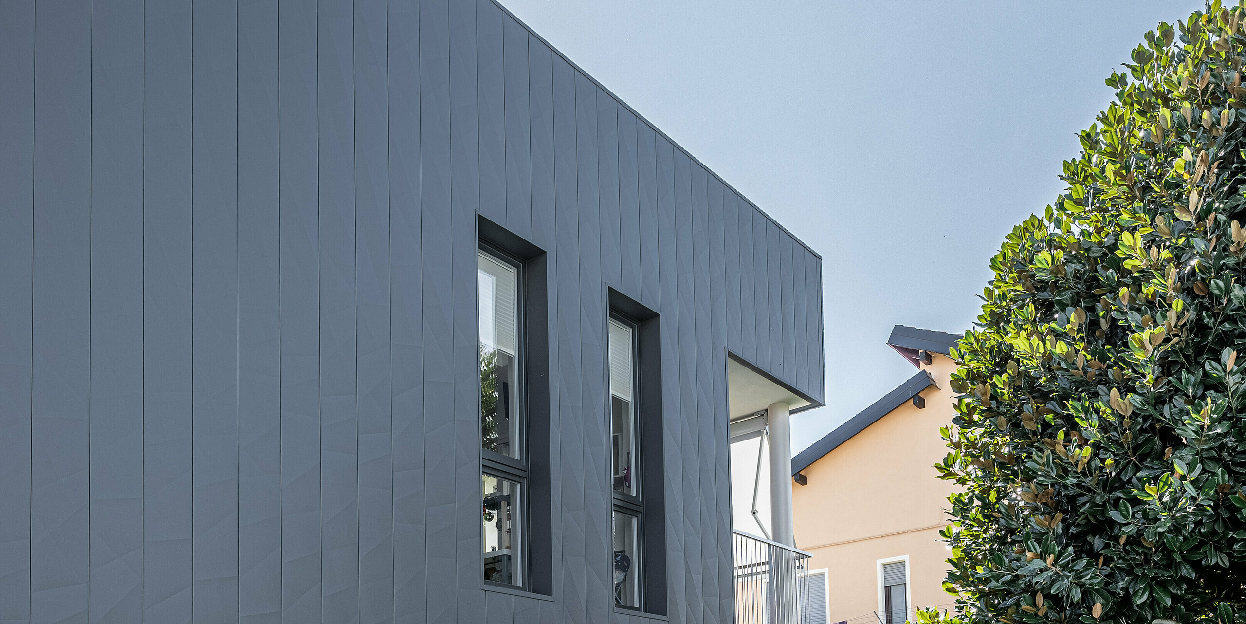 ertikale Ansicht eines zeitgenössischen Einfamilienhauses in Savigliano, Italien, ausgestattet mit einer dunkelgrauen PREFA Siding.X Aluminiumfassade. Die Fassade aus Blech zeichnet sich durch ihre einzigartige matte Oberfläche und geometrische Struktur aus, die das moderne Design des Hauses betont. Großflächige Glasfronten bieten einen transparenten Übergang zwischen dem Innenraum und dem sorgfältig angelegten Garten, wodurch eine offene und einladende Wohnatmosphäre entsteht.