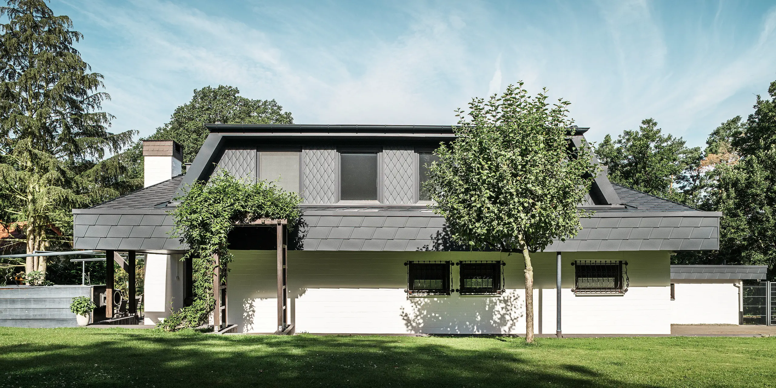 Framifrån sett på trädgårdssidan av en villa i Mechernich med ett stort trapezformat takfönster, täckt med små PREFA-takrombar P.10 antracit. Taket är täckt med PREFA-takshingel DS.19 i samma nyans. Fasader på nedre våningen är vitmålade och en grön pergola fulländar helhetsintrycket i trädgårdsdelen. Träd och buskar ramar in utsikten och skapar en harmonisk integrering i naturen.