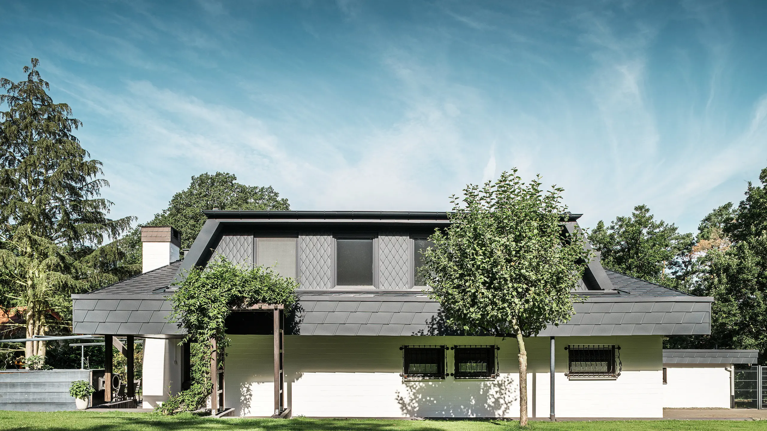 Framifrån sett på trädgårdssidan av en villa i Mechernich med ett stort trapezformat takfönster, täckt med små PREFA-takrombar P.10 antracit. Taket är täckt med PREFA-takshingel DS.19 i samma nyans. Fasader på nedre våningen är vitmålade och en grön pergola fulländar helhetsintrycket i trädgårdsdelen. Träd och buskar ramar in utsikten och skapar en harmonisk integrering i naturen.