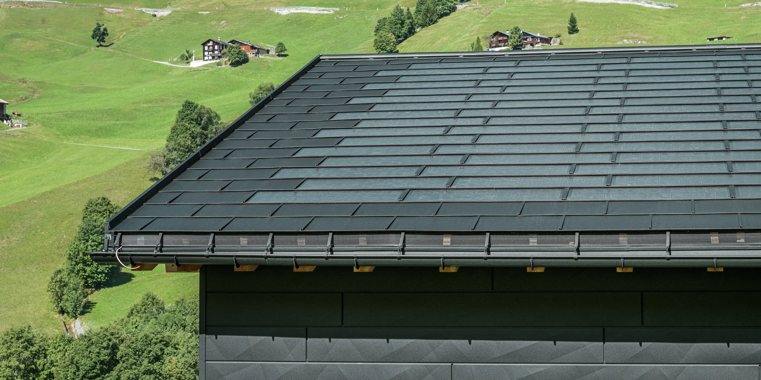 Närbild av takfoten på en modern villa i den alpina omgivningen i Ascharina i Schweiz. Taket har täckts med den innovativa PREFA soltakplattan och den traditionella takplattan R.16, båda i P.10 svart. PREFA hängränna och PREFA avloppsrör säkerställer en tillförlitlig takavvattning. Längst ned på bilden syns de omisskännliga fasadpanelerna PREFA Fasadpanel.X. De rena linjerna hos aluminiumprodukterna understryker den moderna designen mitt i ett pittoreskt bergslandskap.