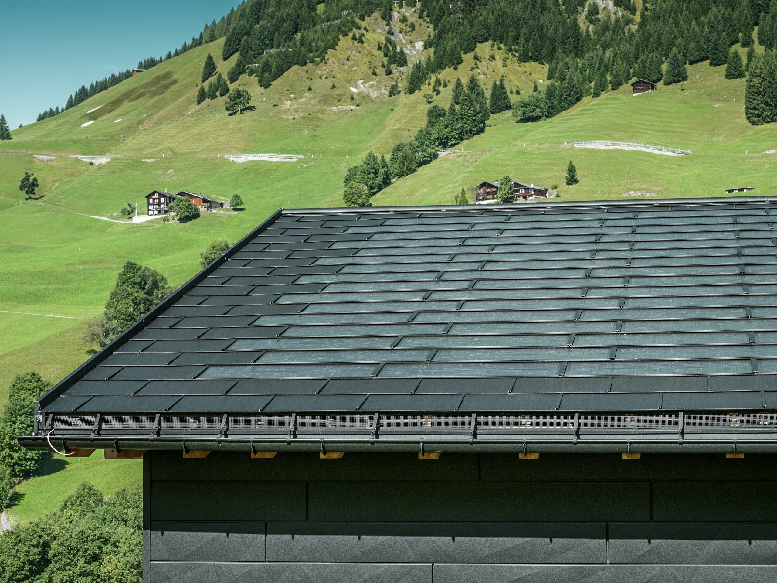 Närbild av takfoten på en modern villa i den alpina omgivningen i Ascharina i Schweiz. Taket har täckts med den innovativa PREFA soltakplattan och den traditionella takplattan R.16, båda i P.10 svart. PREFA hängränna och PREFA avloppsrör säkerställer en tillförlitlig takavvattning. Längst ned på bilden syns de omisskännliga fasadpanelerna PREFA Fasadpanel.X. De rena linjerna hos aluminiumprodukterna understryker den moderna designen mitt i ett pittoreskt bergslandskap.