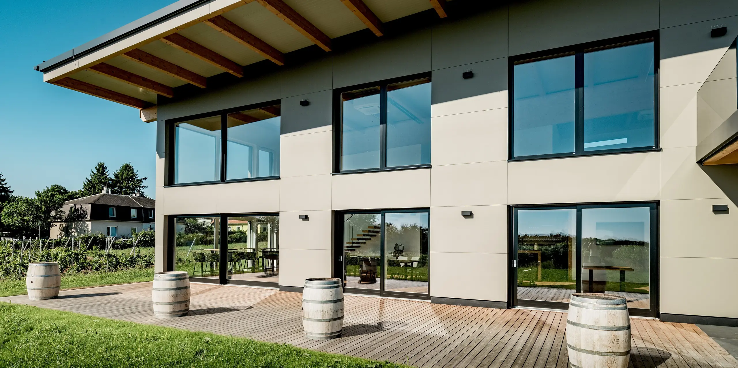 Frontale Ansicht des Weinguts Nigl in Perchtoldsdorf mit moderner Aluminiumfassade von PREFA in P.10 Prefabronze. Die glatte Oberfläche der Verbundplatten und die symmetrische Fensteranordnung prägen das klare, geometrische Fassadenbild. Große Glasflächen reflektieren den Himmel und bieten spannende Einblicke ins lichtdurchflutete Innere mit sichtbarer Treppe. Der darüberliegende Dachvorsprung mit offenen Holzbalken sorgt für natürlichen Sonnenschutz. Im Vordergrund runden Weinbarriquefässer und eine großzügige Holzterrasse die Szene ab. Die PREFA Aluminium Verbundplatte steht hier für moderne Optik, hohe Wetterbeständigkeit und langfristige Farbtreue.