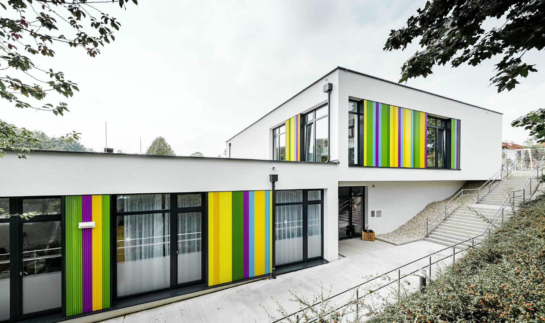 Färgglada fasadkomponenter från PREFA för dagiset i Hargelsberg. Byggnaden har ett plant tak och stora fönster från golv till tak.