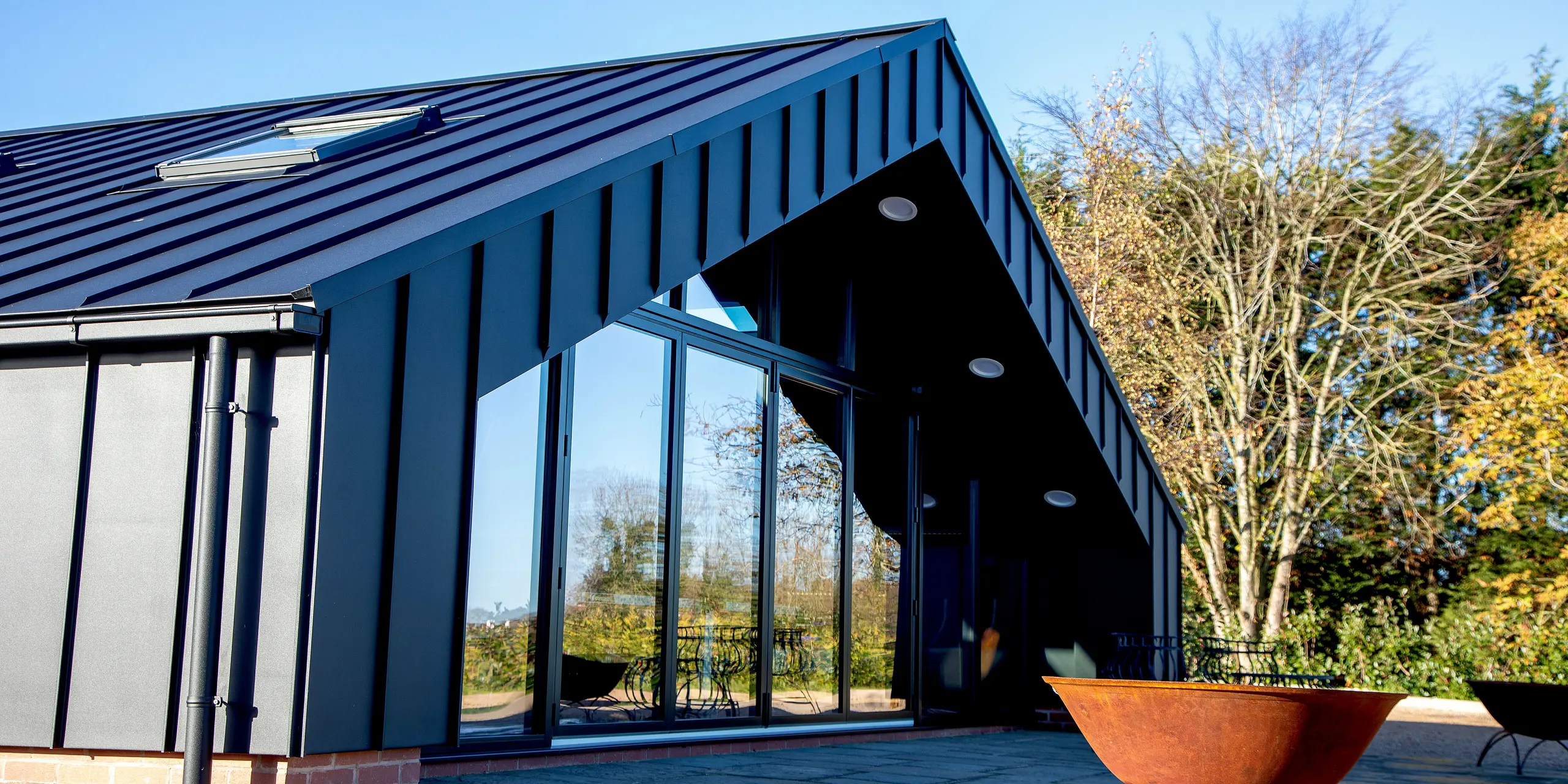 Froschperspektive auf das moderne Freizeitgebäude mit vollständig verkleideter PREFA Aluminiumfassade und PREFALZ Dachfläche in P.10 Schwarz. Die markanten Stehfalzlinien, die klare Dachgeometrie und die großzügige Glasfront erzeugen eine kraftvolle architektonische Präsenz. Die witterungsbeständige Aluminiumausführung garantiert höchste Stabilität und langlebigen Schutz im Außenbereich.
