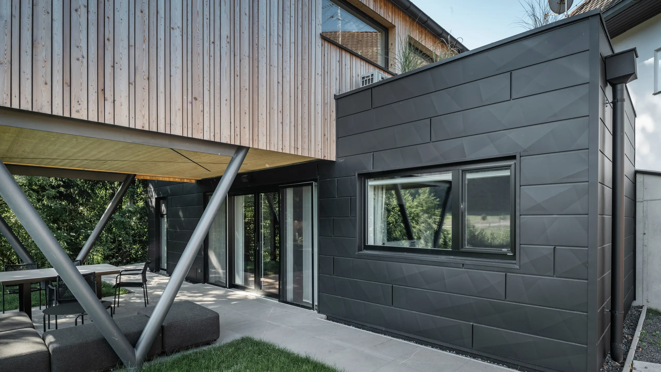 Arkitektoniskt höjdpunkt: ”Woodstock Chalet 5” i St. Martin am Innkreis, Oberösterreich. Den moderna bröllopslokalen framhävs av användningen av antracitfärgade PREFA Fasadpanel.X. Aluminiumelementen bildar en stark kontrast till de ljusa träpanelerna och de metalliska stödkonstruktionerna. De mörka fönsterramarna smälter harmoniskt in i fasadens utformning, vilket ger byggnaden ett elegant utseende. Denna bild visar den estetiska balansen mellan naturlig trästruktur och den industriella charmen hos aluminiumfasaden, som står för utformningen av moderna byggnader.