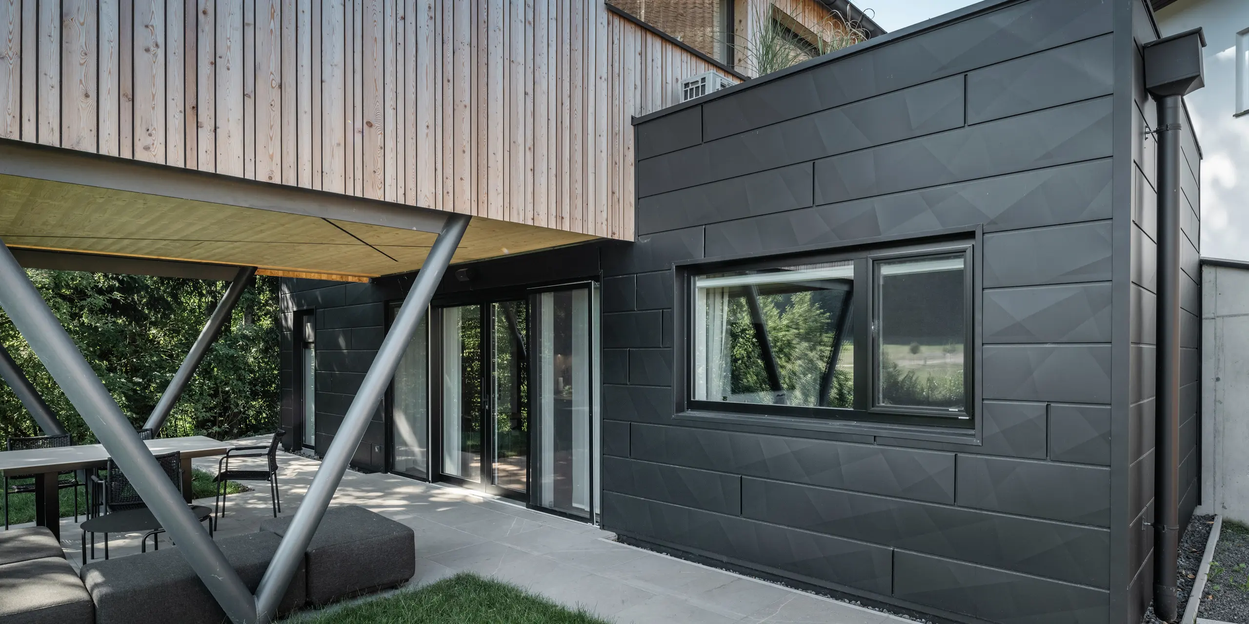 Arkitektoniskt höjdpunkt: ”Woodstock Chalet 5” i St. Martin am Innkreis, Oberösterreich. Den moderna bröllopslokalen framhävs av användningen av antracitfärgade PREFA Fasadpanel.X. Aluminiumelementen bildar en stark kontrast till de ljusa träpanelerna och de metalliska stödkonstruktionerna. De mörka fönsterramarna smälter harmoniskt in i fasadens utformning, vilket ger byggnaden ett elegant utseende. Denna bild visar den estetiska balansen mellan naturlig trästruktur och den industriella charmen hos aluminiumfasaden, som står för utformningen av moderna byggnader.