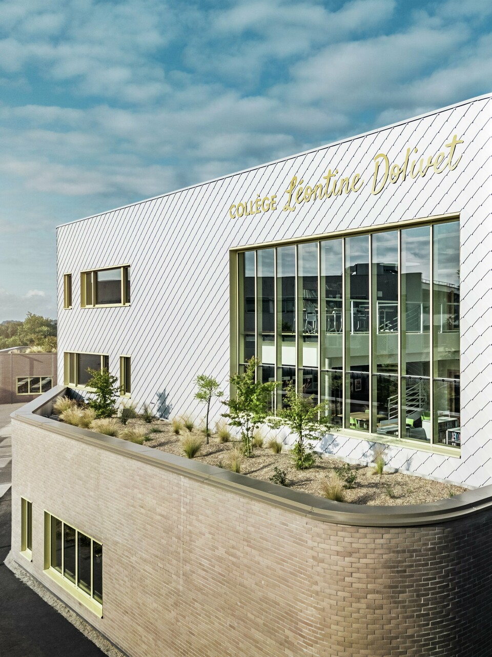 Das Foto zeigt eine markante Ecke des Collège Léontine Dolivet in Cesson-Sévigné. Die Fassade besteht aus silbermetallischen PREFA-Wandelementen, die diagonal angeordnet sind und das Licht reflektieren. Besonders auffällig ist die geschwungene Form des Obergeschosses, die dem Gebäude eine sanfte und moderne Anmutung verleiht. Große Glasflächen geben den Blick ins Innere frei, während gelbe Fensterrahmen farbliche Akzente setzen. Im Vordergrund befindet sich eine bepflanzte Fläche mit Kies und Sträuchern, die die Architektur harmonisch ergänzt. Der leicht bewölkte Himmel hebt die Metalloberfläche zusätzlich hervor.