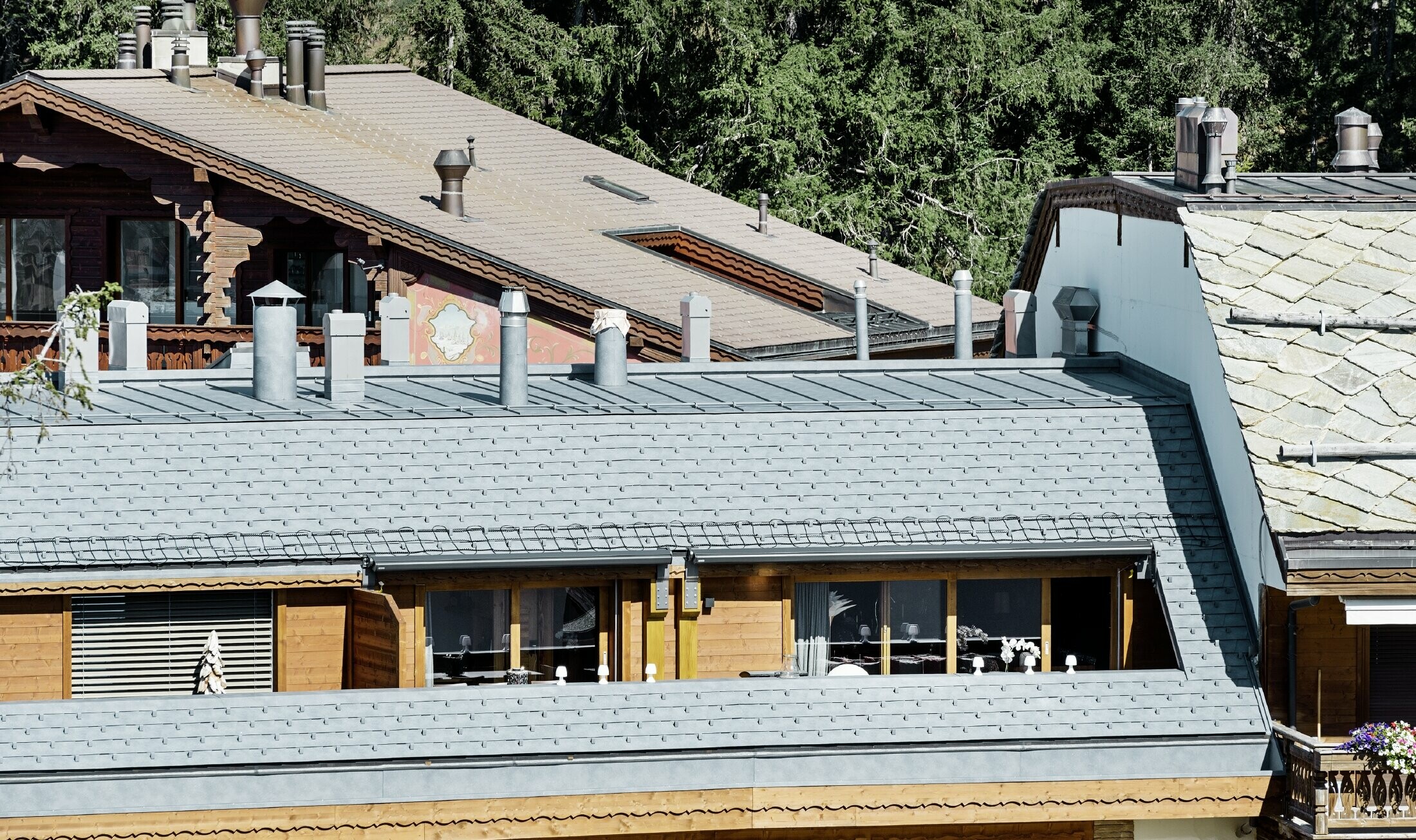Flerfamiljshus i Crans-Montana med utsikt över bergen och en fasad med lekfulla träelement med ett tak av PREFA:s takshingel i stengrå aluminium