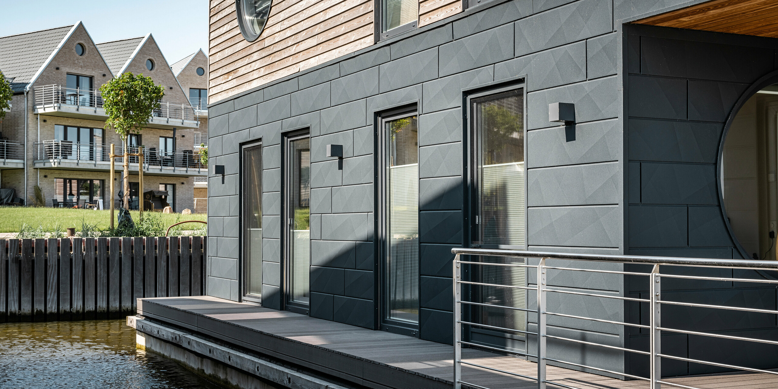 Detailaufnahme eines Wasserhauses im Pionierhafen Schleswig, das die hochwertige PREFA Siding.X Fassade in P.10 Anthrazit zeigt. Das Zusammenspiel von moderner Aluminiumverkleidung und natürlichen Holzelementen spiegelt eine Symbiose aus Funktionalität und Design wider. Im Hintergrund ergänzen traditionelle Häuser mit Giebeldächern und begrünten Außenbereichen die Szenerie und betonen das harmonische Zusammenspiel von alter und neuer Architektur am Wasser.